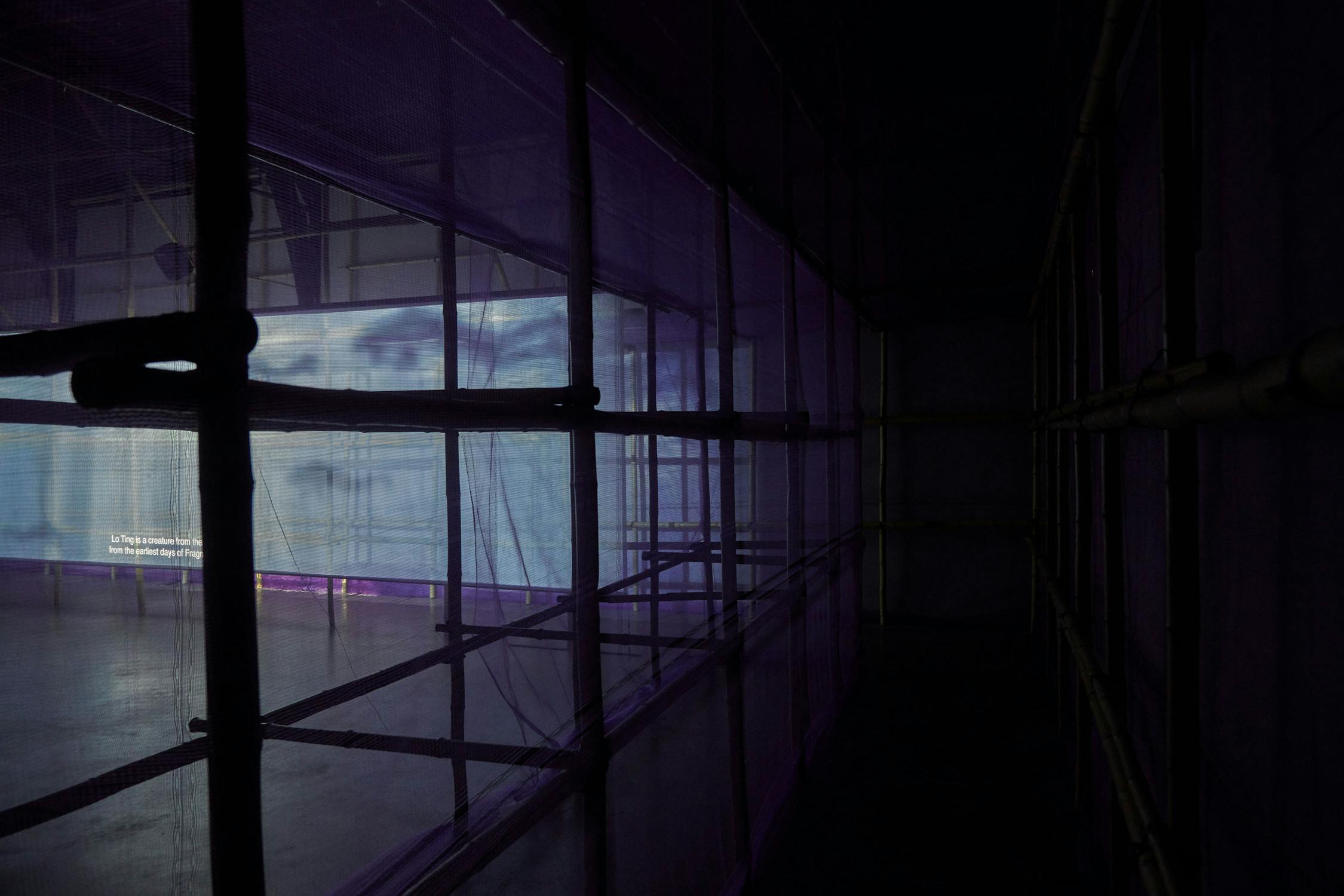 A dimly lit room features a large installation with mesh screens and metal scaffolding. The left side is illuminated with a bluish light, and a section of white text is visible on the distant wall, providing contrast against the dark surroundings.