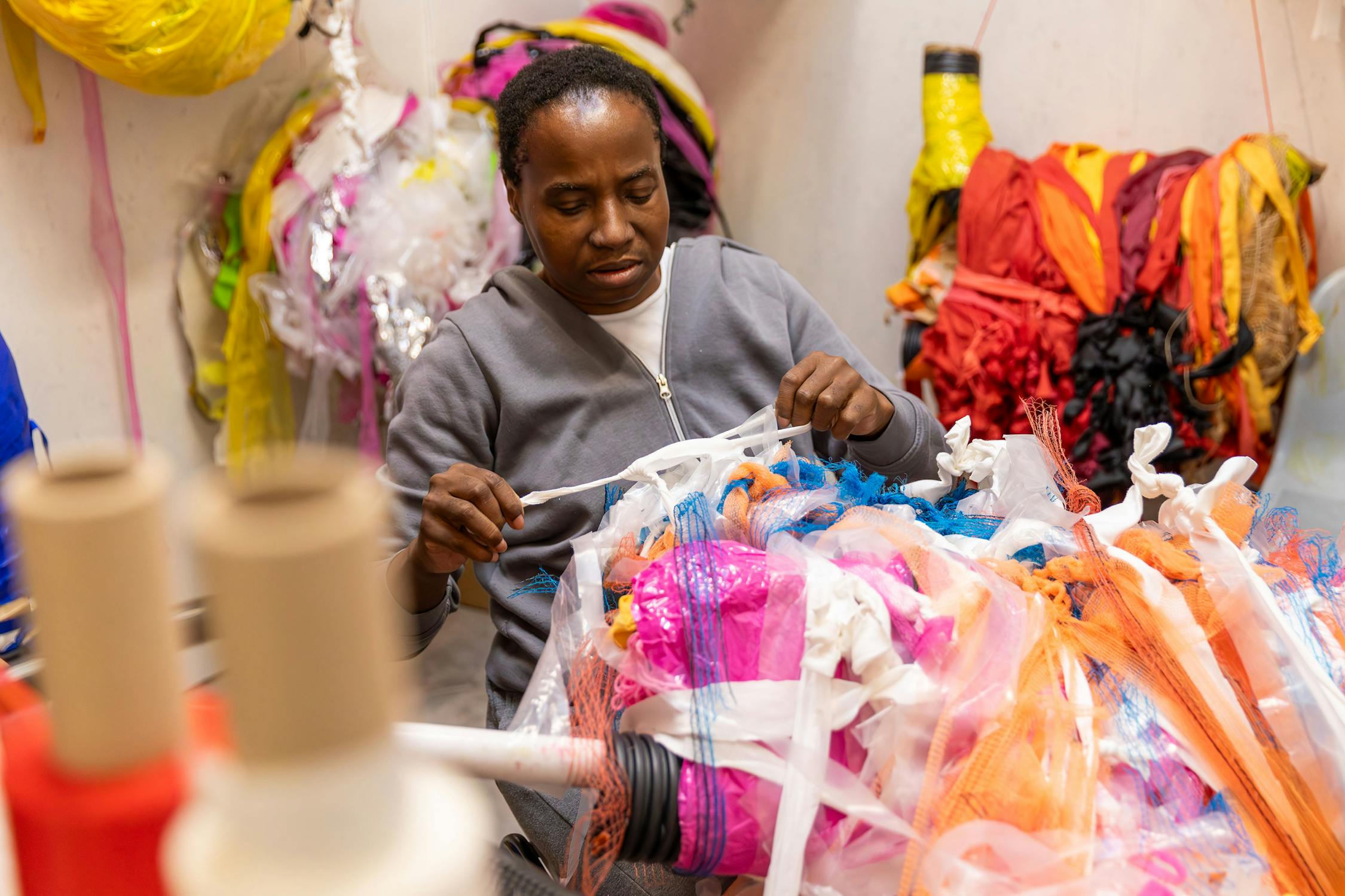 Artist Nnena Kalu carefully inspects vibrant, multicoloured plastic materials in her studio. Shelves and bins around them are filled with similar colourful materials. Industrial spools are visible in the foreground.