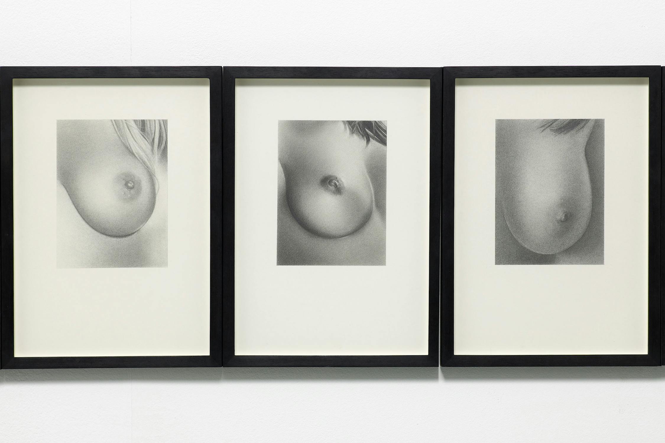 Three framed graphite drawings of breasts installed on a white wall.