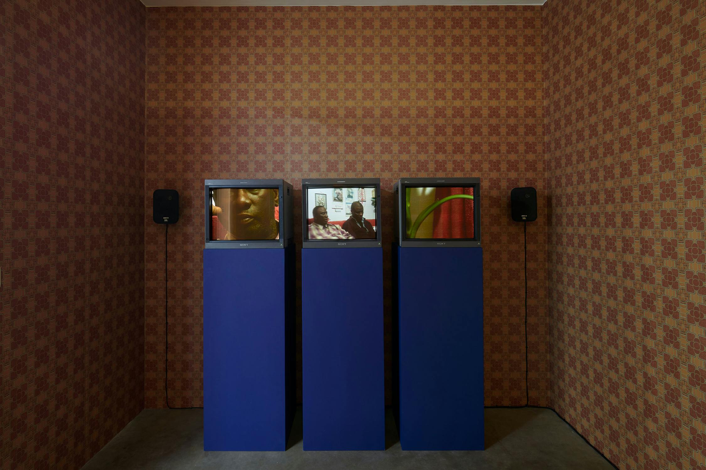 Three blue plinths each supporting an old TV, set against a wall with vintage patterned brown and orange wallpaper. The screens display various close ups of people. 