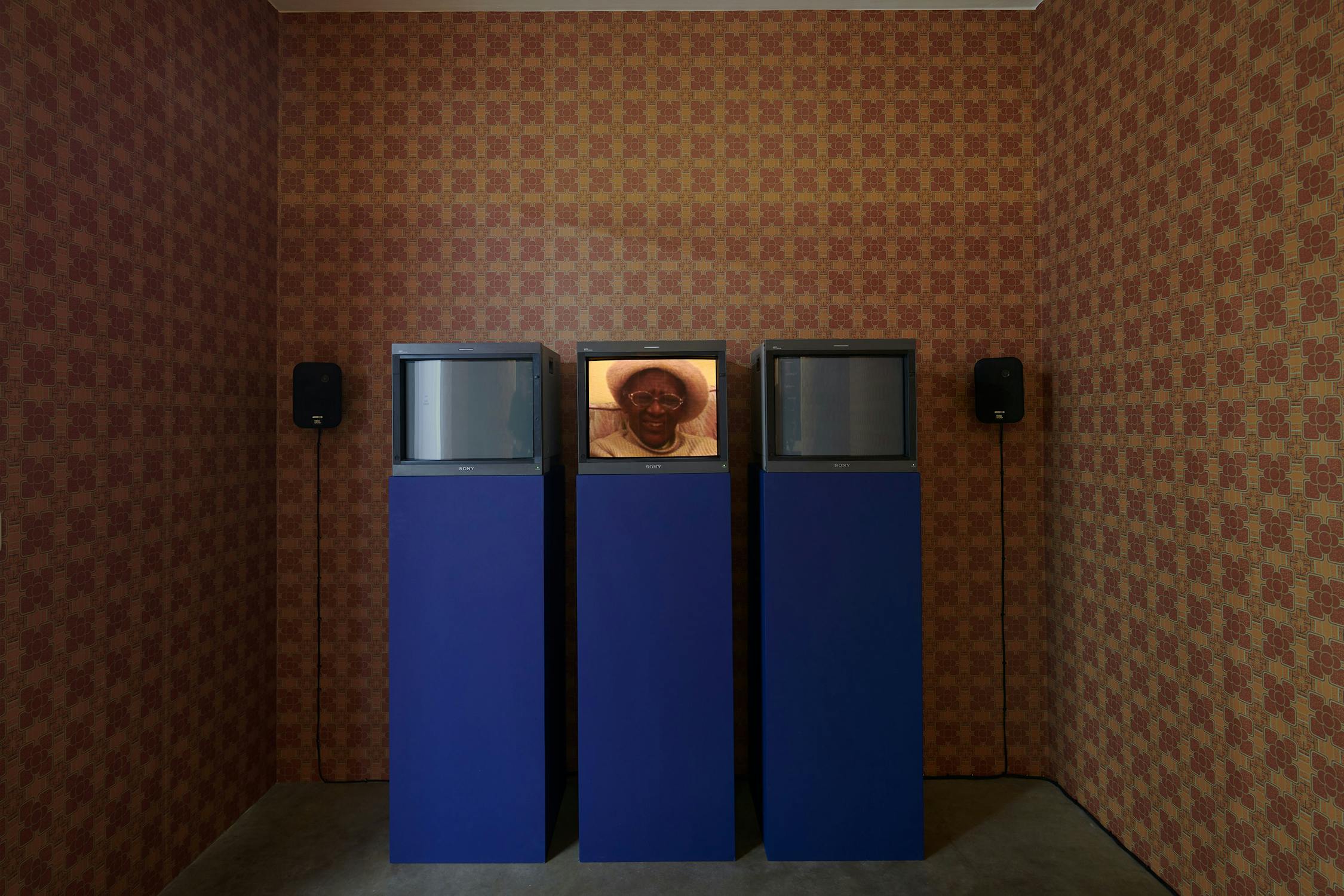 Three blue plinths each supporting an old TV, set against a wall with vintage patterned brown and orange wallpaper. The screens display various close ups of people.