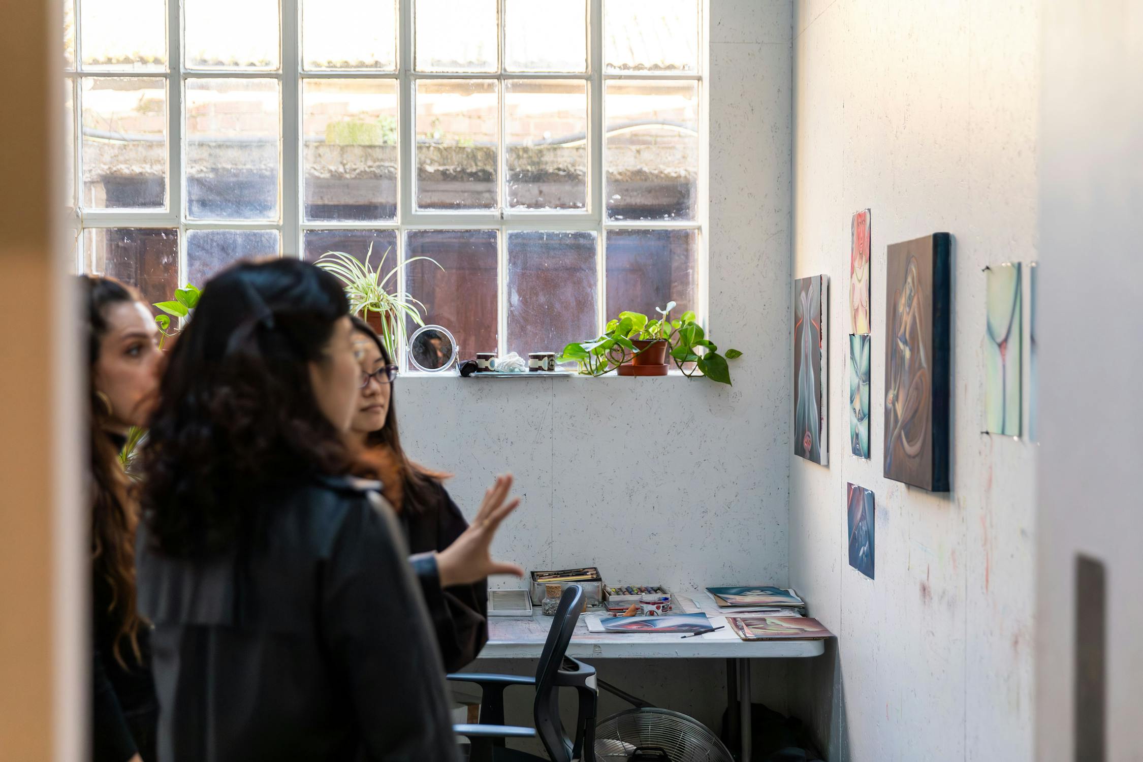 Three people are observing artwork displayed on a white wall in a small room with a large window. There are some potted plants and various art supplies on a shelf by the window. The room is well lit with natural light coming through the window.