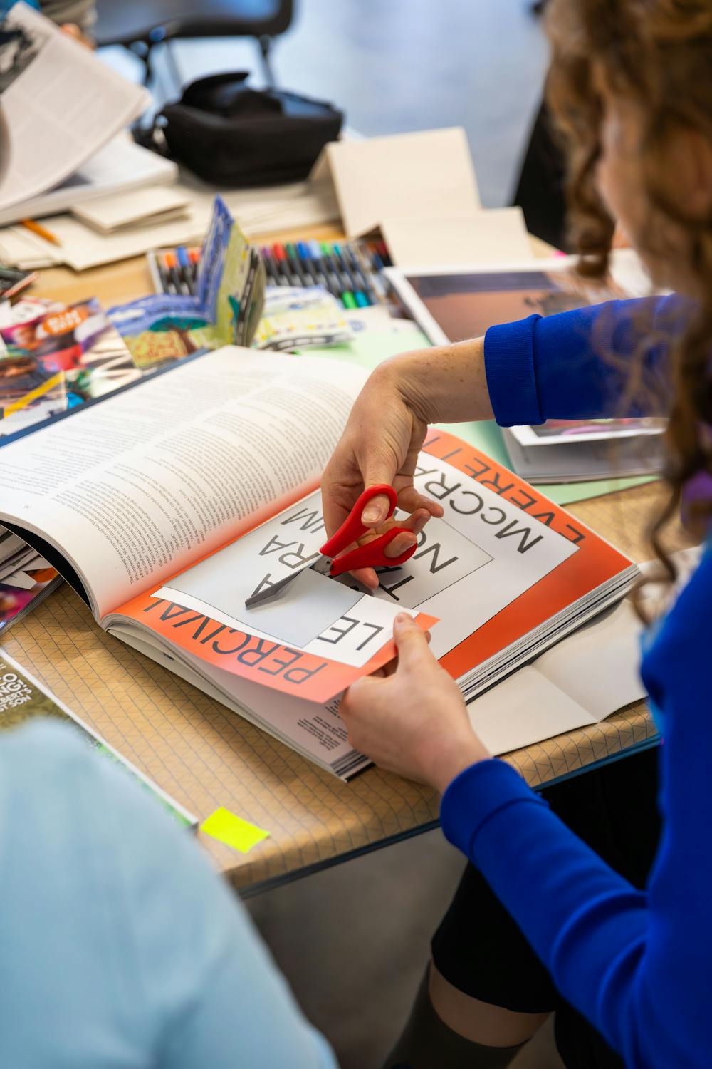 A person wearing a blue shirt is using red scissors to cut out a page from an open book on a cluttered table. Various art supplies and paper materials are scattered around, indicating a crafting or design project in progress.