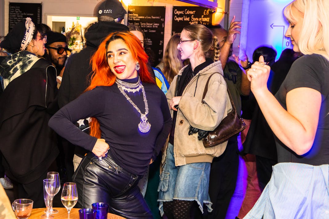 A woman with vibrant red hair and wearing a black top and leather pants is smiling and posing with one hand on her hip at a lively party. Others around her are chatting and dancing. Various drinks are visible on a table in the foreground.