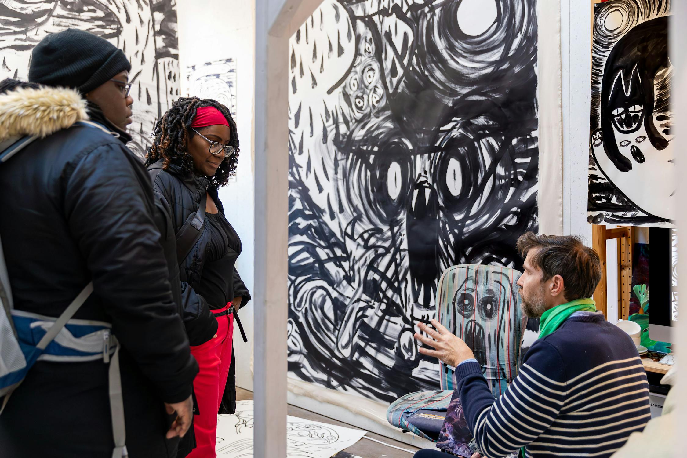 A person in a striped shirt and green scarf sits and gestures while talking to two standing individuals in a room decorated with large, dark, expressive art pieces on the walls. The standing individuals wear coats and hats. The setting appears to be an art studio or gallery.