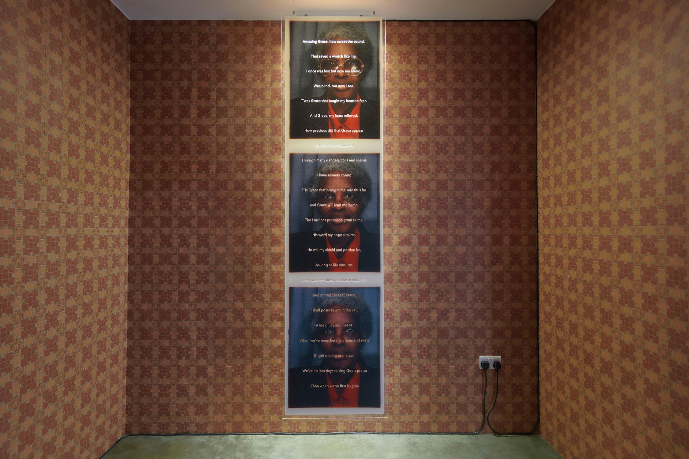 An exhibition display features four vertically aligned framed posters with text overlaying images of a person. The posters are suspended from the ceiling in a room with vintage patterned wallpapered.