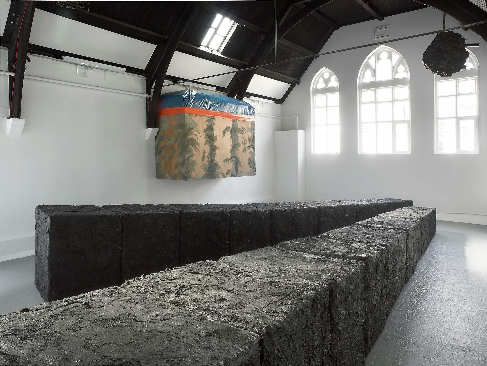A contemporary art installation by Phyllida Barlow in a bright room with large arched windows. Black rectangular blocks form a zigzag pattern on the floor. A textured wall hanging with a nature-inspired design is displayed near the ceiling.