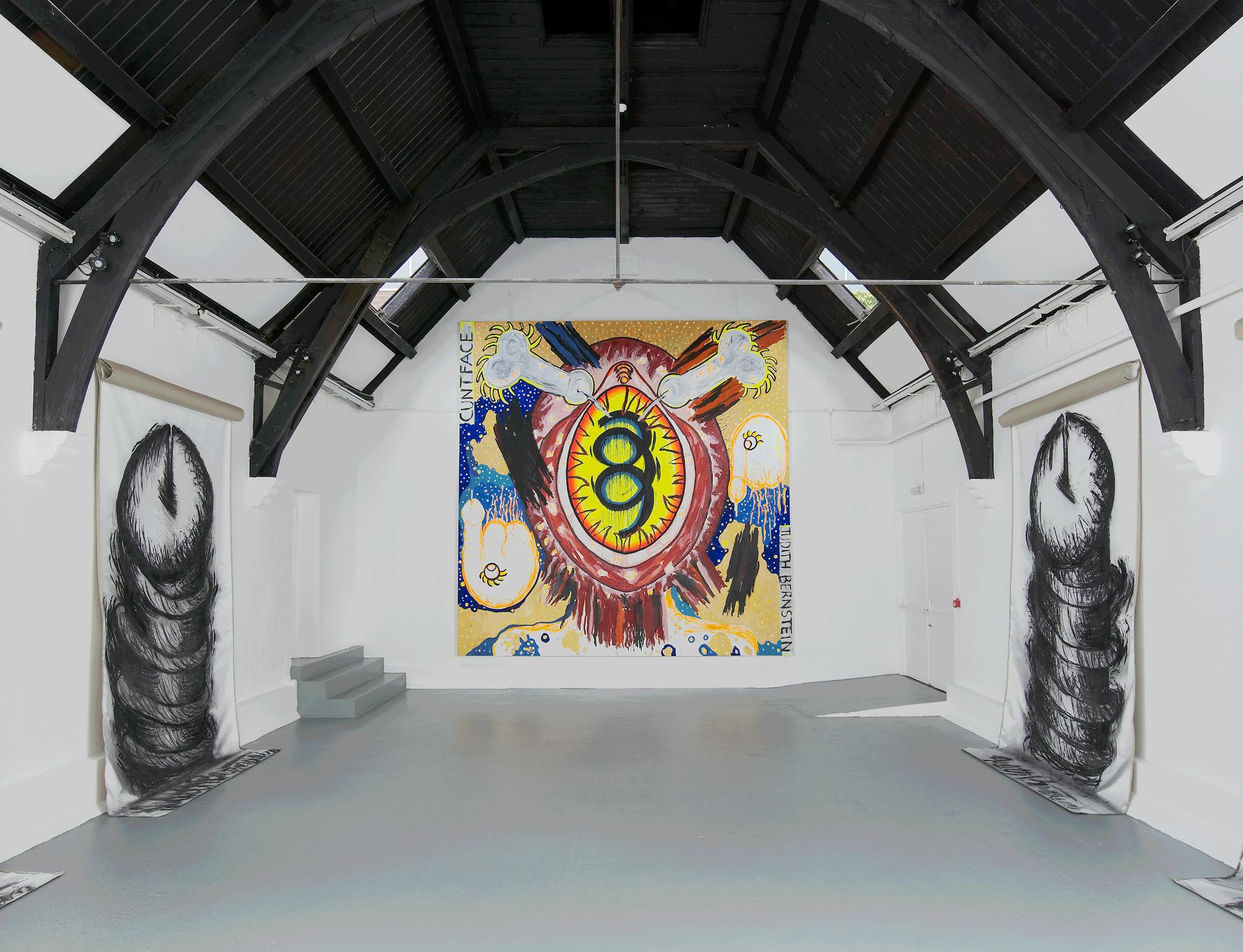 An art gallery room with a high, arched ceiling. Works by Judith Bernstein, including a large, vibrant painting featuring a cosmic vagina with surrounding patterns is on the far wall. Two black and white phallic abstract artworks are hung on either side. The room is lit with overhead lights.