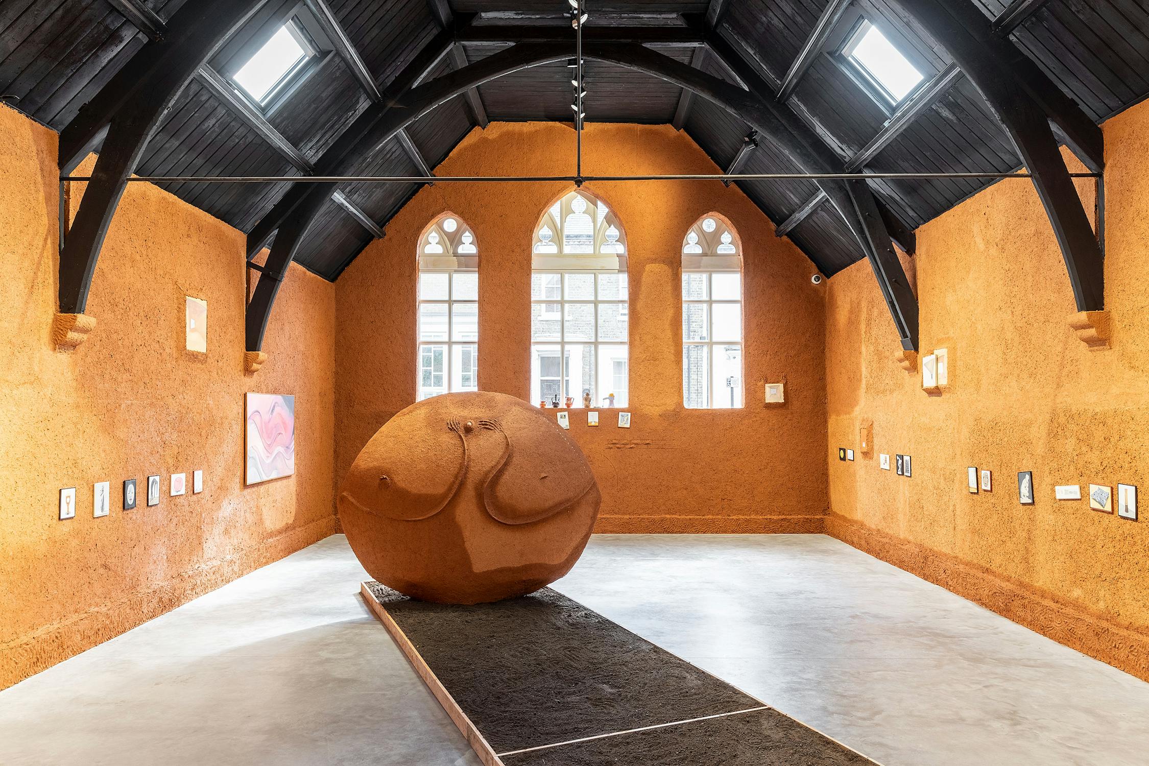 An art gallery room with orange adobe walls and a high, dark arched ceiling featuring large gothic windows. In the centre is a large, round sculpture. Paintings and small artworks are displayed on the walls.