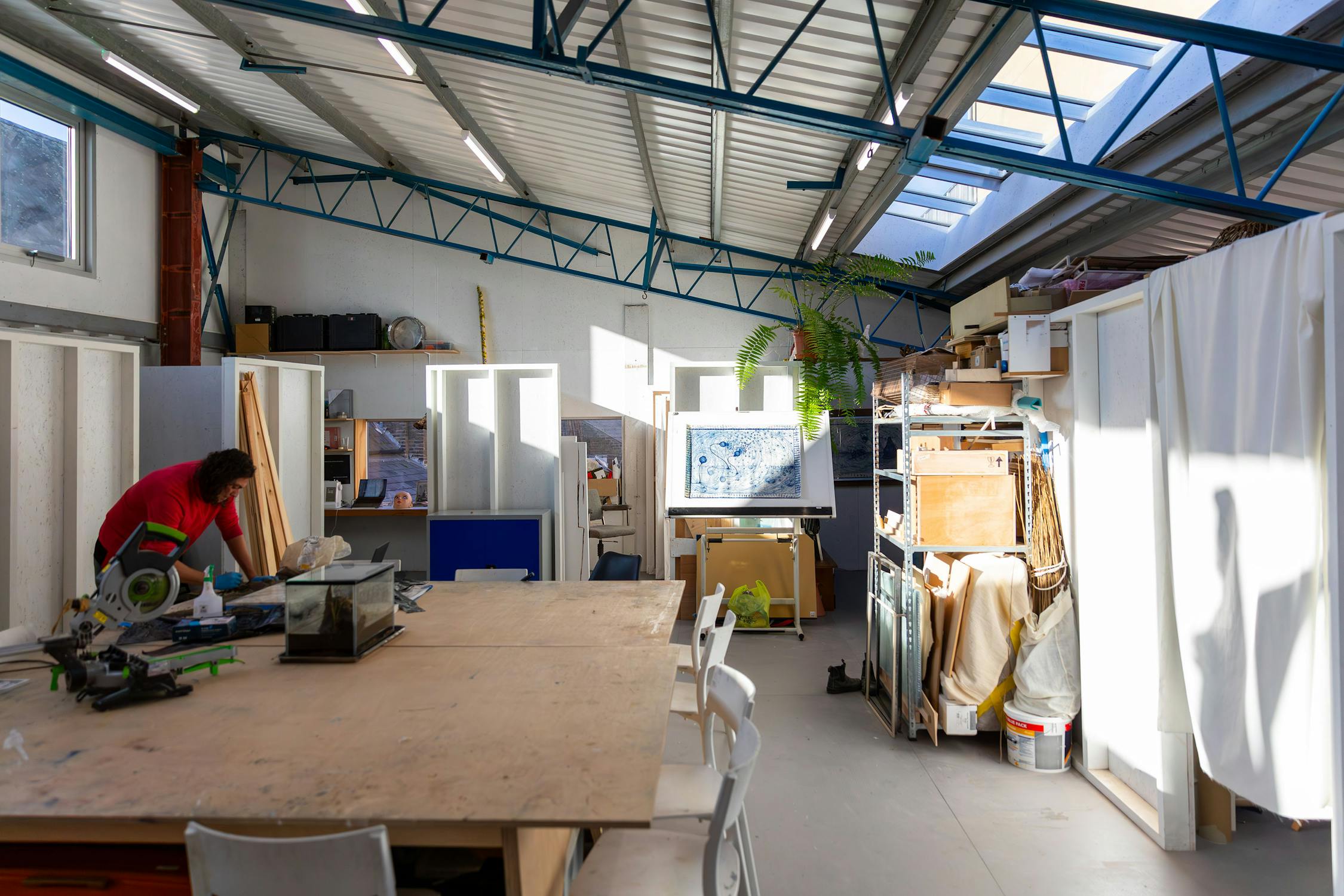 A well-lit art studio with large windows and a high ceiling. A person works at a cluttered desk on the left. An art piece is displayed on an easel nearby, surrounded by various supplies and furniture.