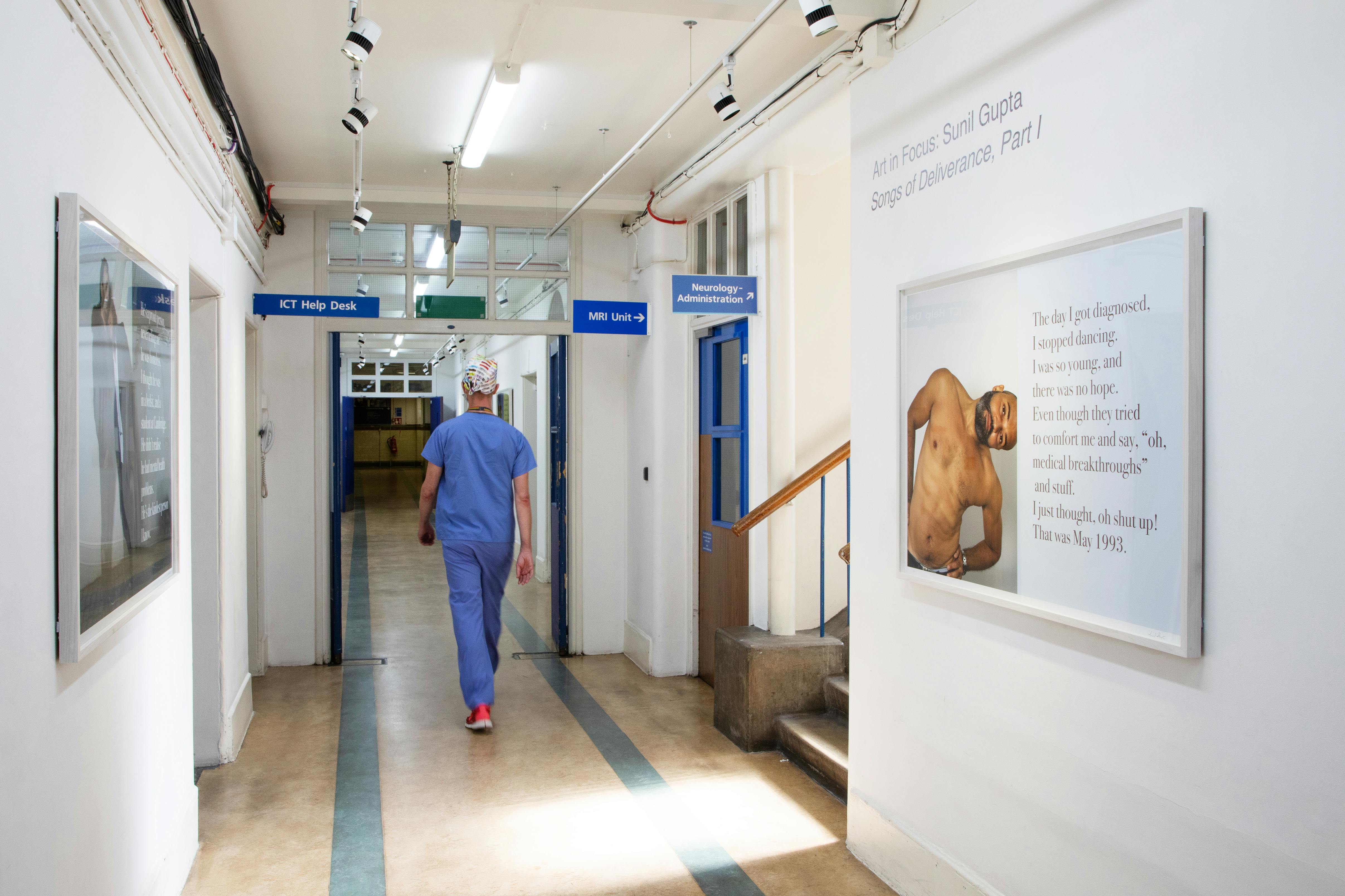 A healthcare worker in blue scrubs walks down a well-lit hospital corridor. The hallway has white walls, framed artwork by Sunil Gupta, and signs indicating the ICT Help Desk and Neurology Administration. Steps lead to a door on the right.