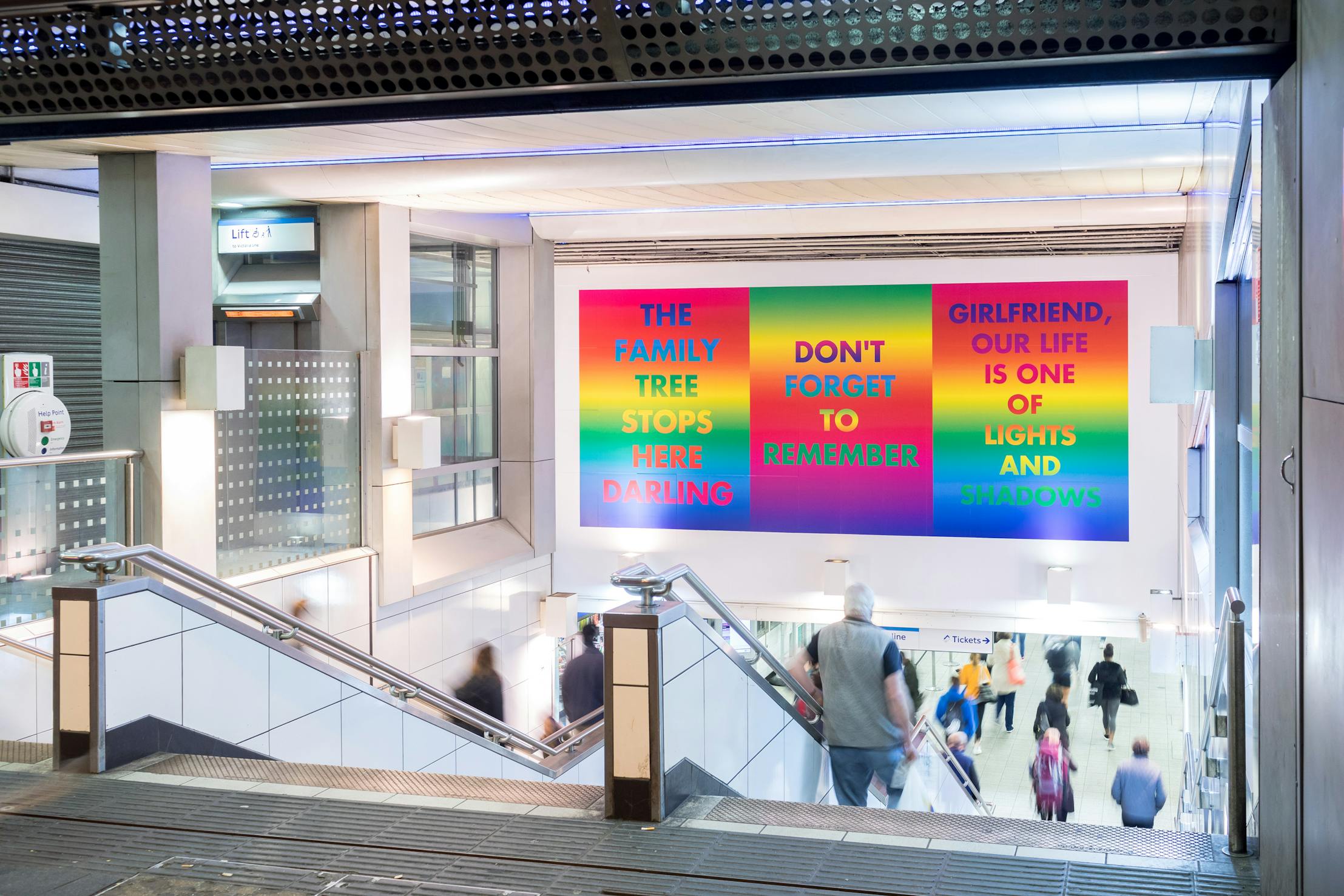 A staircase in a Brixton Underground station leads down to people walking. Three colourful wall posters by David McDiarmid display text: "THE FAMILY TREE STOPS HERE DARLING," "DON’T FORGET TO REMEMBER," and "GIRLFRIEND, OUR LIFE IS ONE OF LIGHTS AND SHADOWS.