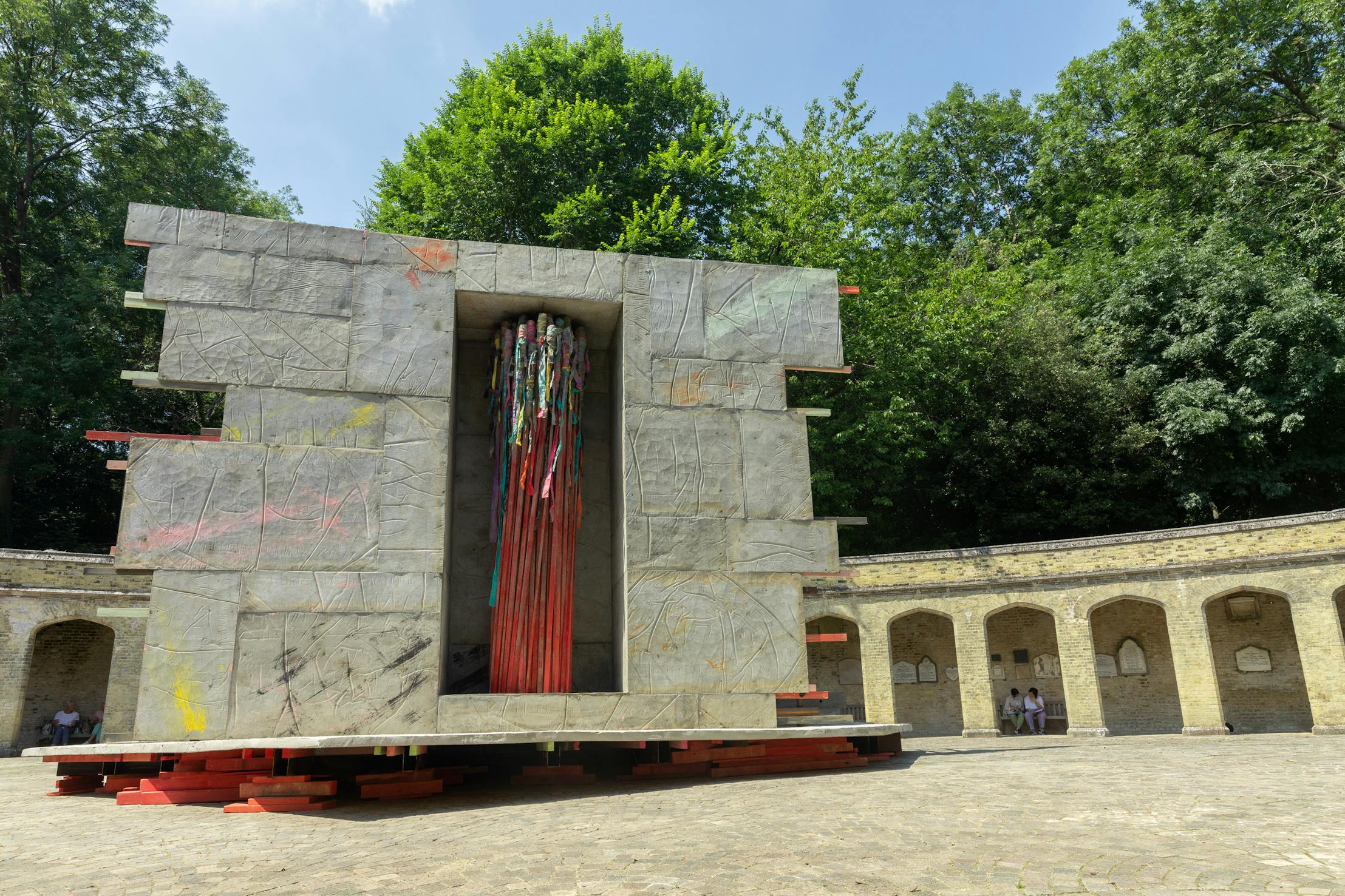 A large, abstract stone and metal sculpture by Phyllida Barlow stands outdoors, featuring colourful vertical rods at its centre. It's set in a circular courtyard surrounded by a stone wall with arched niches. Lush green trees frame the background under a clear sky.