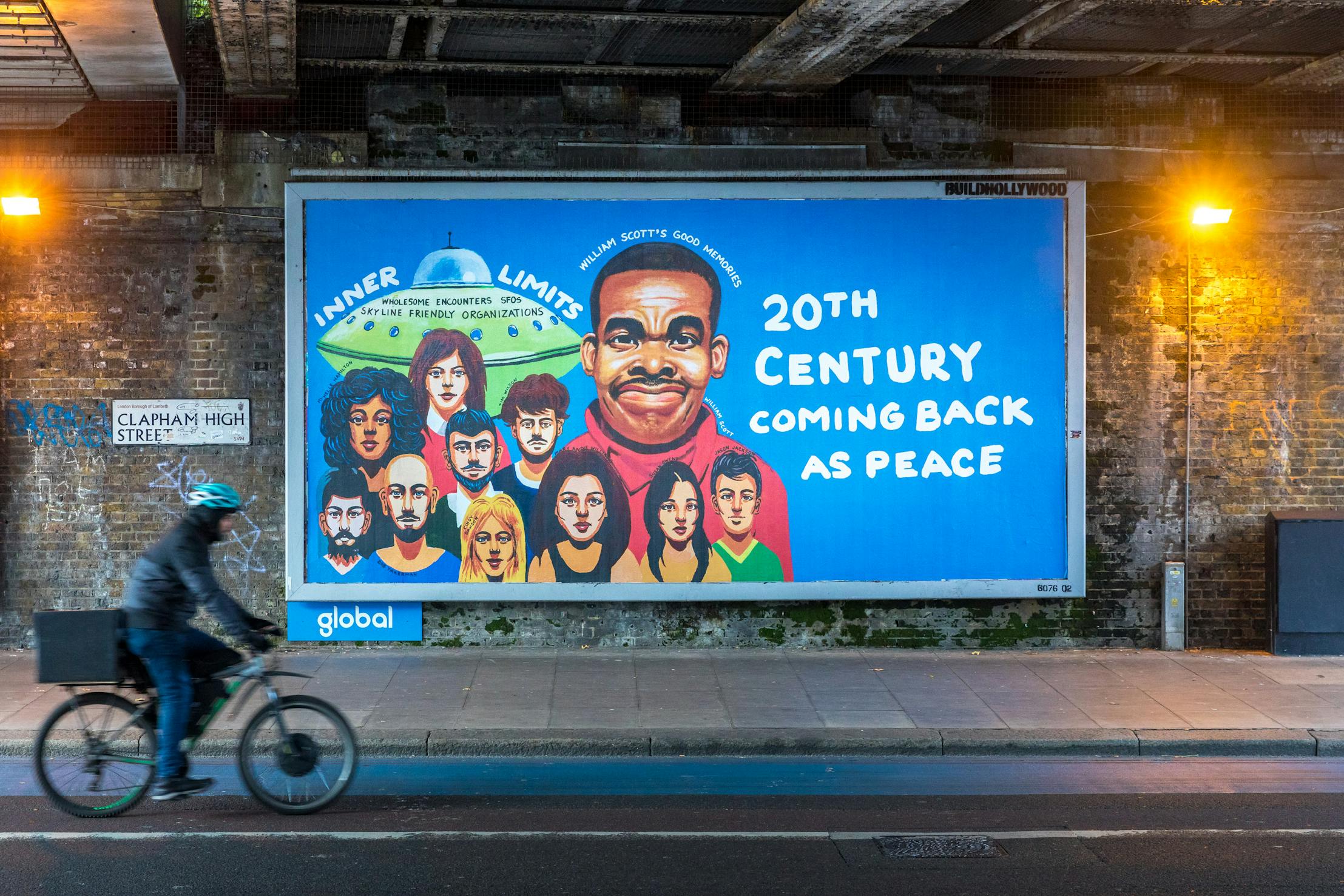 A cyclist rides past a colourful mural by William Scott on a brick wall under a bridge. The mural features a group of diverse individuals beneath a prominent portrait of a man, with text reading "20th Century Coming Back as Peace" and "Inner Limits" in bold letters.