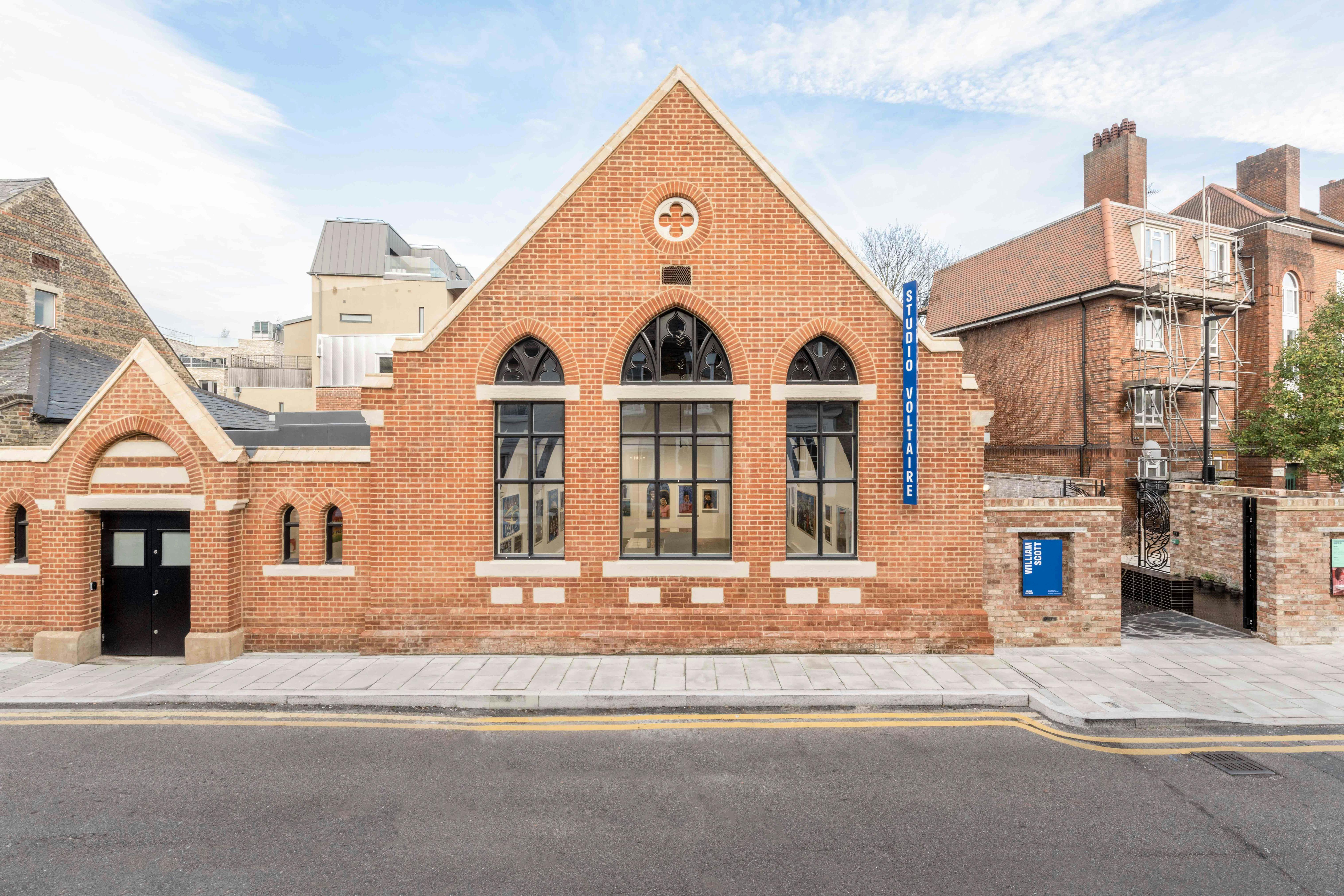 The red-brick exterior of Studio Voltaire, with arched windows and a triangular roof, displaying a blue "Studio Voltaire" sign. The street in front is empty, and several other buildings are visible in the background under a partly cloudy sky.