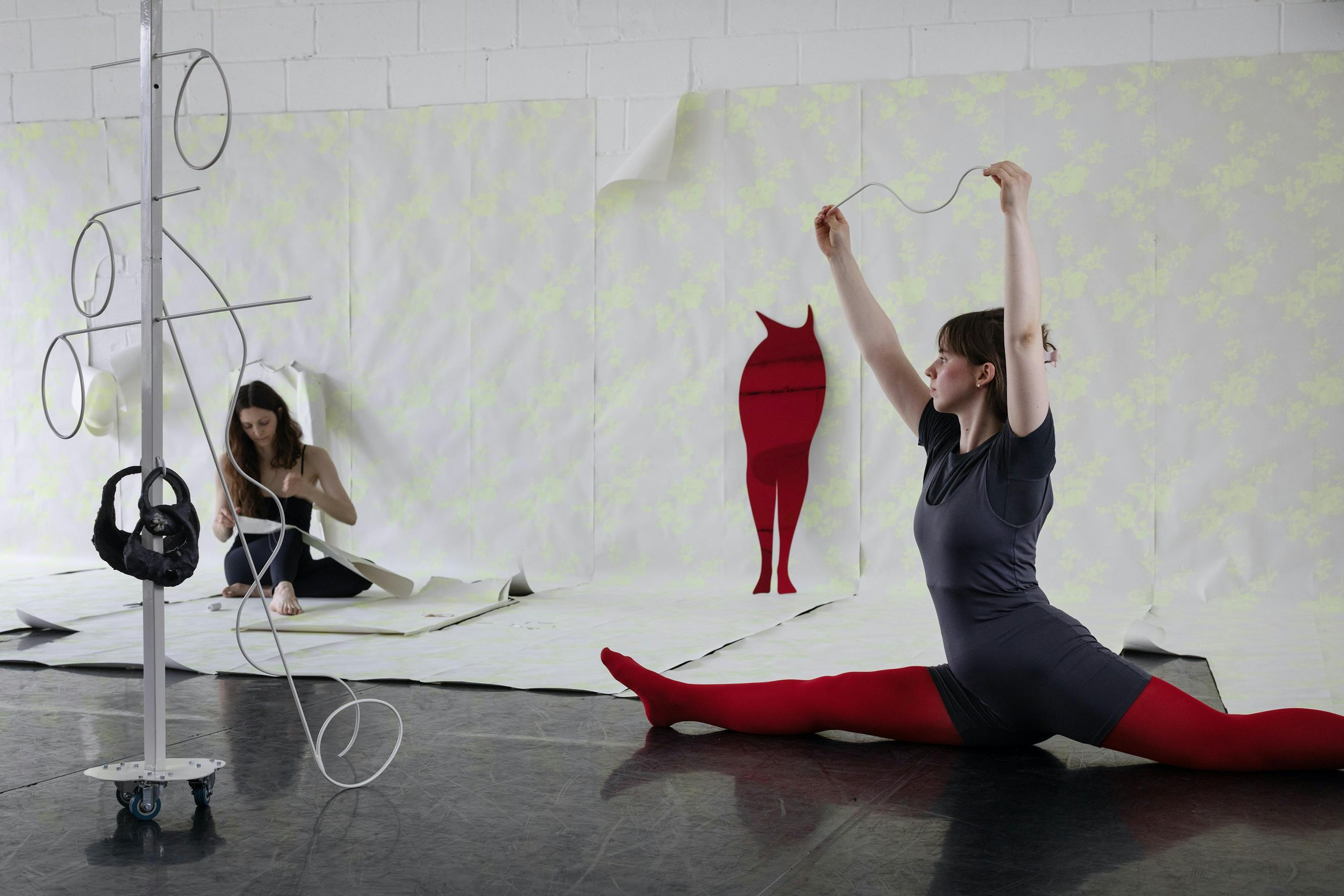 Two women in a studio; one is stretching in a split while holding up string, wearing red tights and a dark outfit. The other is seated on the floor, working with paper. A metal stand is in the foreground, and a red silhouette decorates the wall.