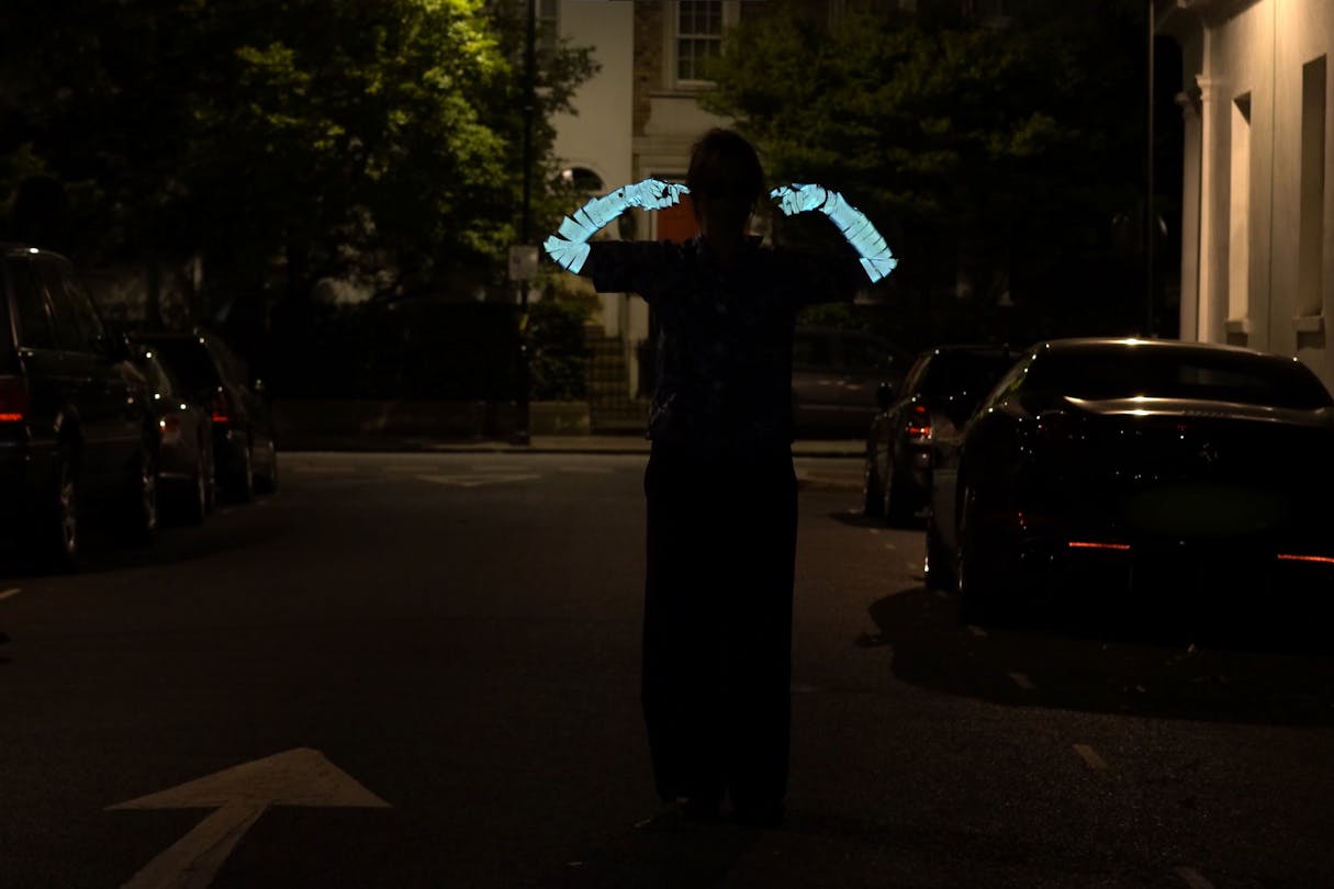 A person stands in the middle of a dimly lit street at night, lifting their arms. The reflective stripes on the sleeves glow under the streetlights. Cars are parked along the street, and trees flank the scene in the background.