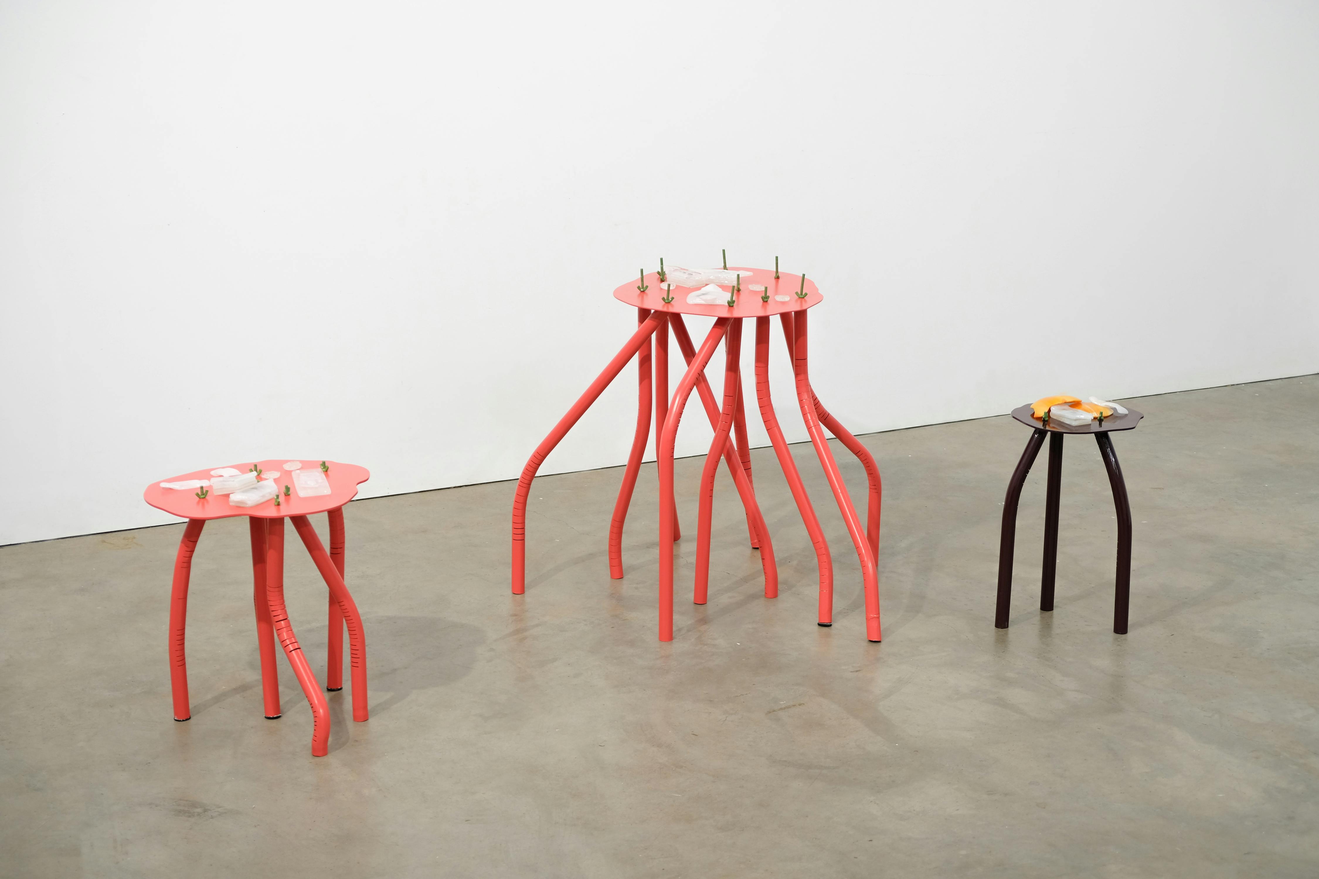 There are three uniquely designed tables with wavy legs: two are coral-coloured with sculptures on top, and one is black with a small object. They stand on a smooth, light-coloured floor against a white wall.