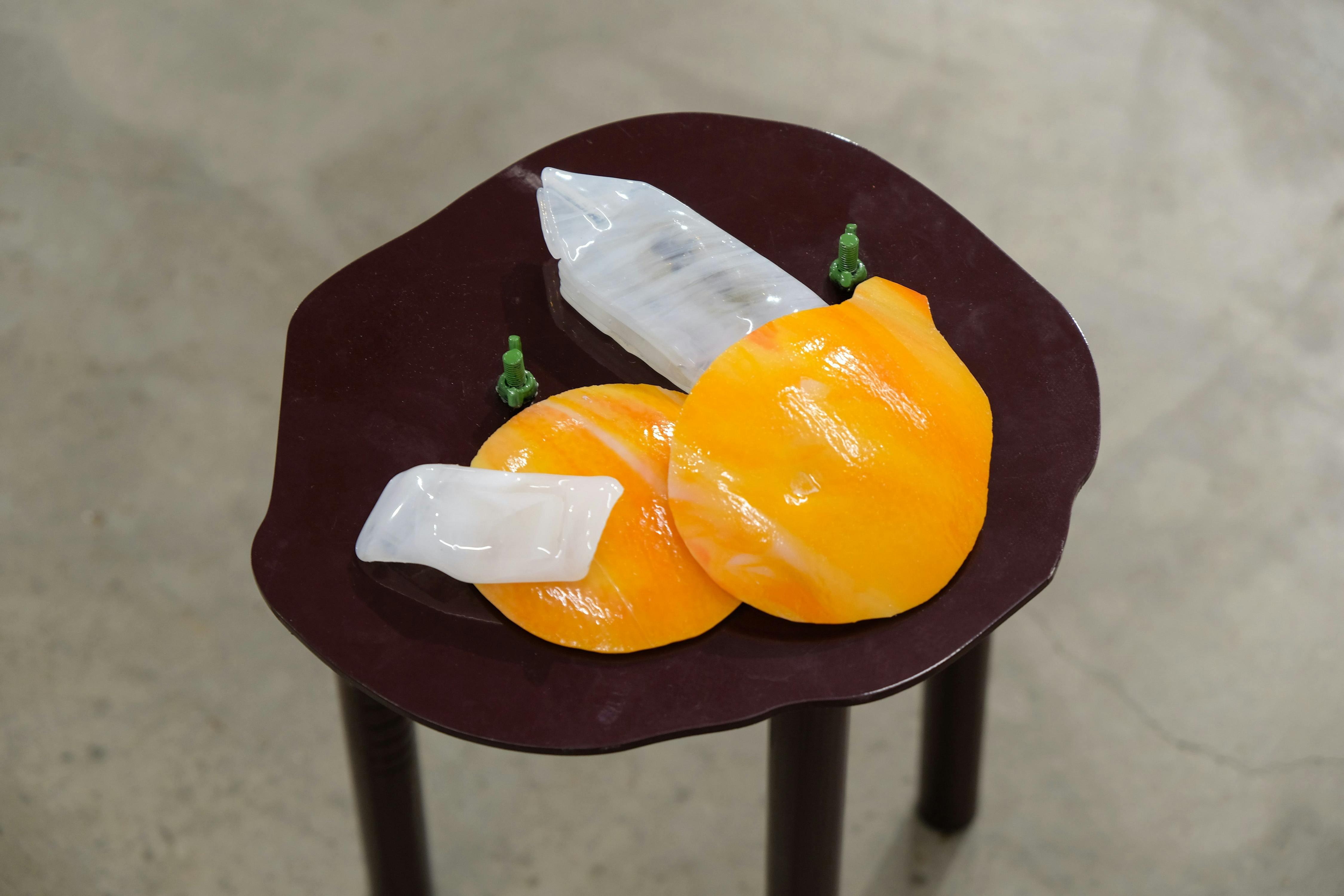 A small stool displays abstract sculptures resembling two orange, circular shapes. The background is a grey concrete floor.