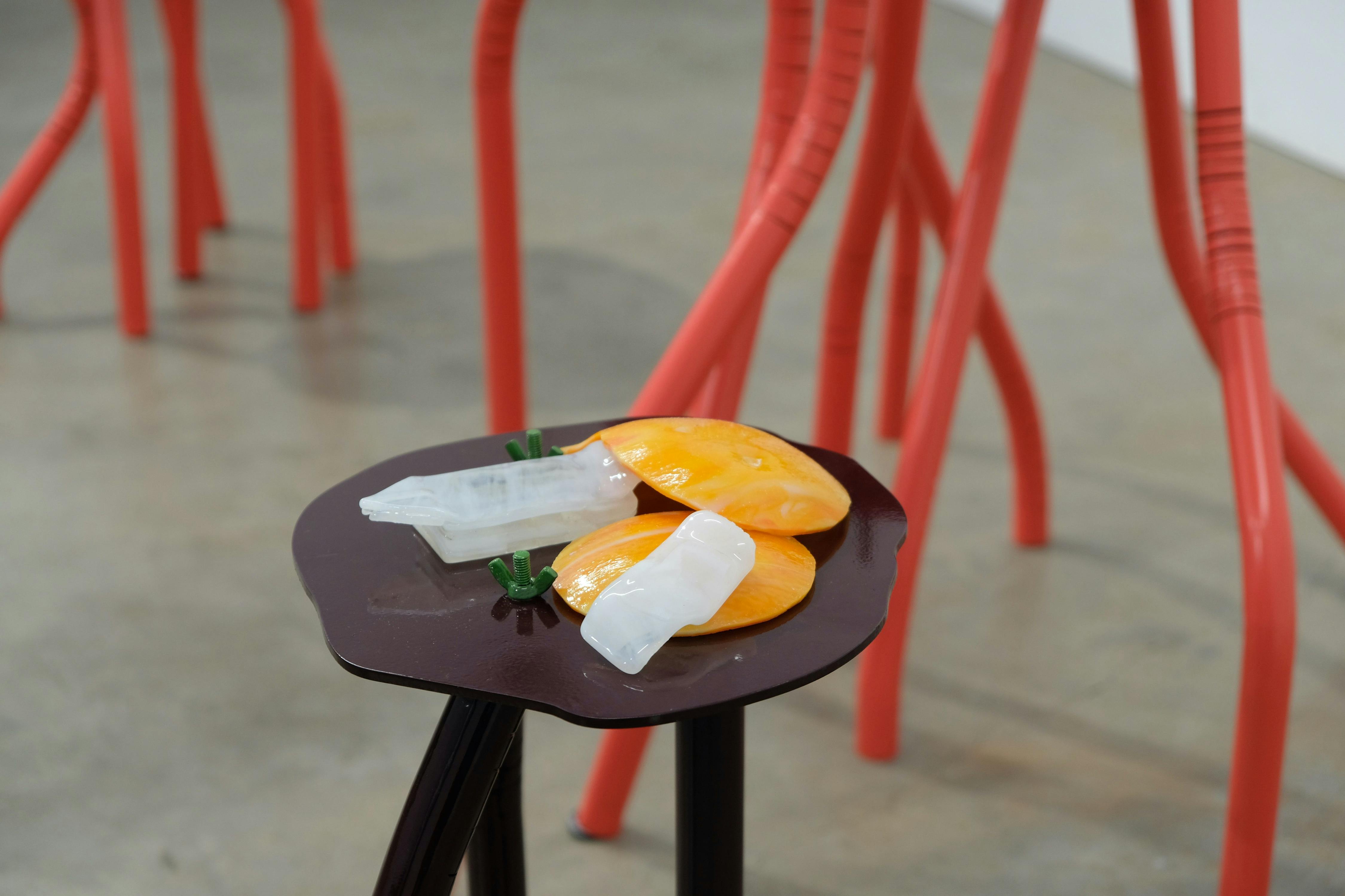 A brown stool holds art resembling a flattened orange and a translucent object. In the background, red, elongated, leg-like sculptures stand on a grey concrete floor.