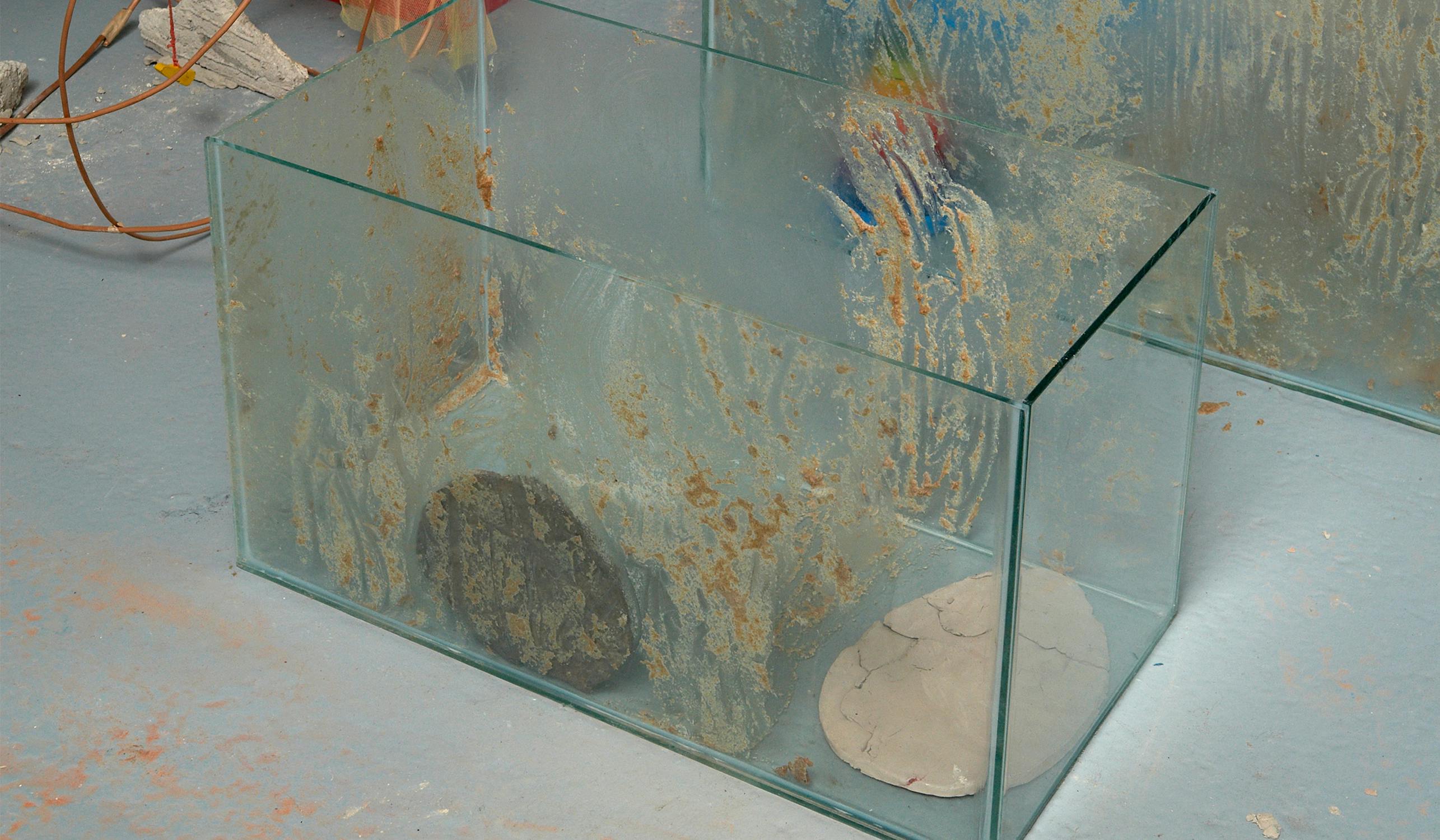 A glass tank with a reflective surface holds a black and a white stone. The tank is surrounded by a lightly dusted floor and is near glass panels with streaks of paint and grime. Wires and a cracked wall are visible in the background.