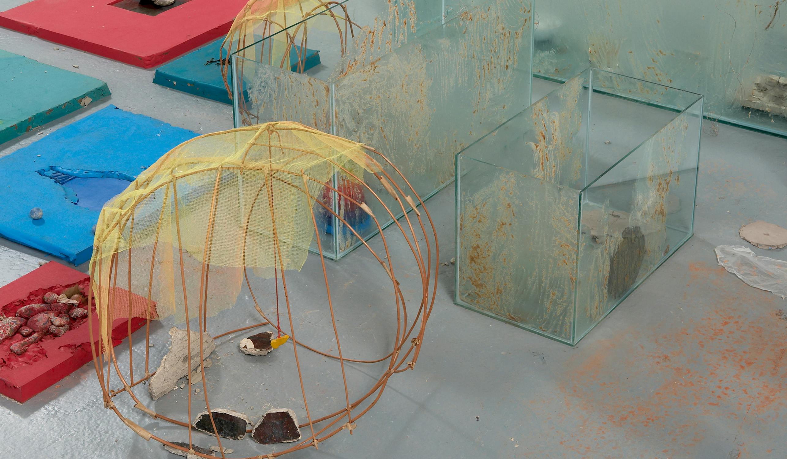 An abstract art installation featuring wireframes, glass boxes, and colorful panels on the floor. The scene includes a yellow mesh-covered dome with scattered objects, creating an eclectic, modern aesthetic.