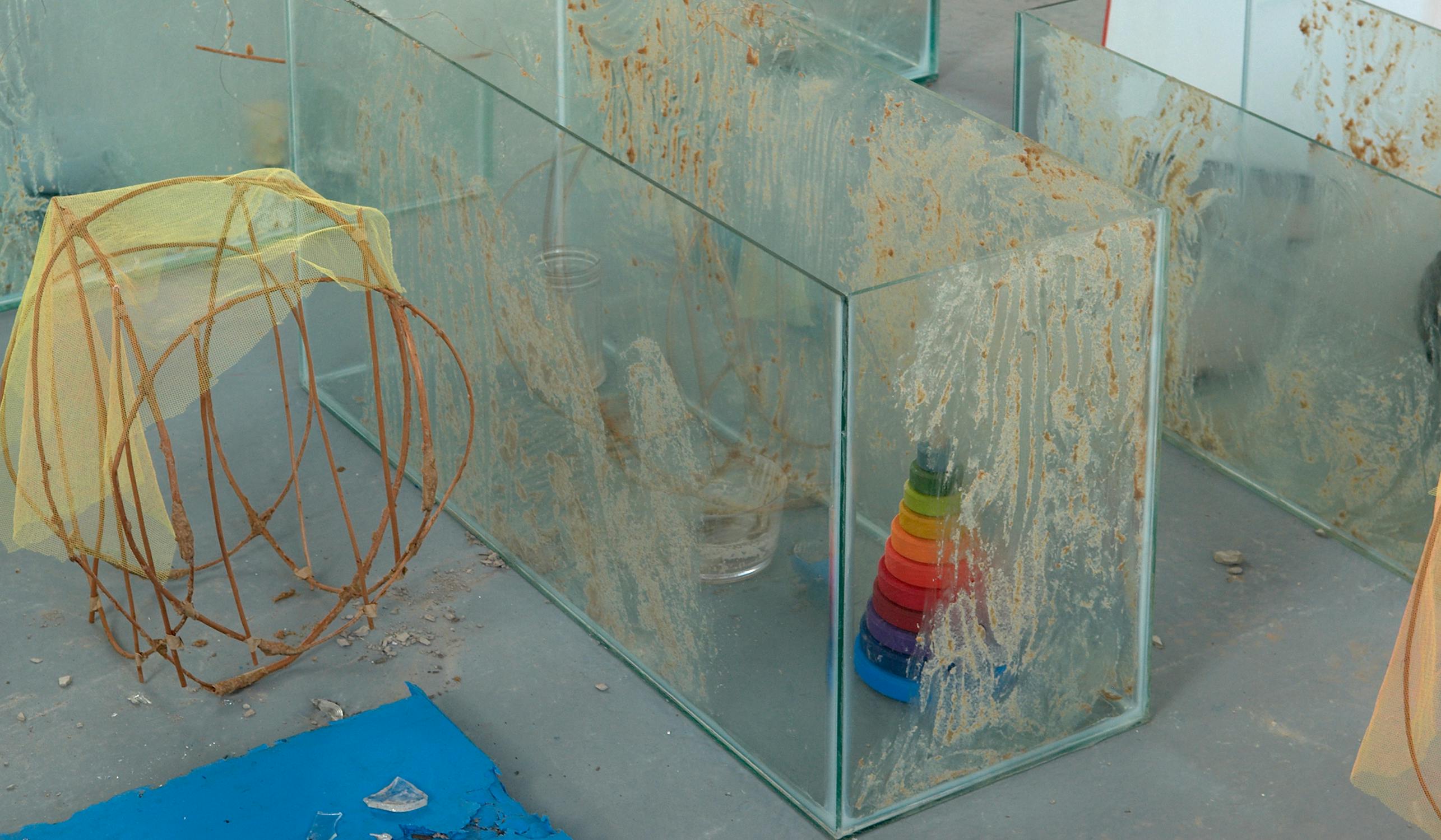 A display of glass boxes, one containing a colorful stackable toy ring. Nearby are yellow fabric and wire structures. The boxes have a textured, dirt-like substance on them. The scene appears abstract and artistic.