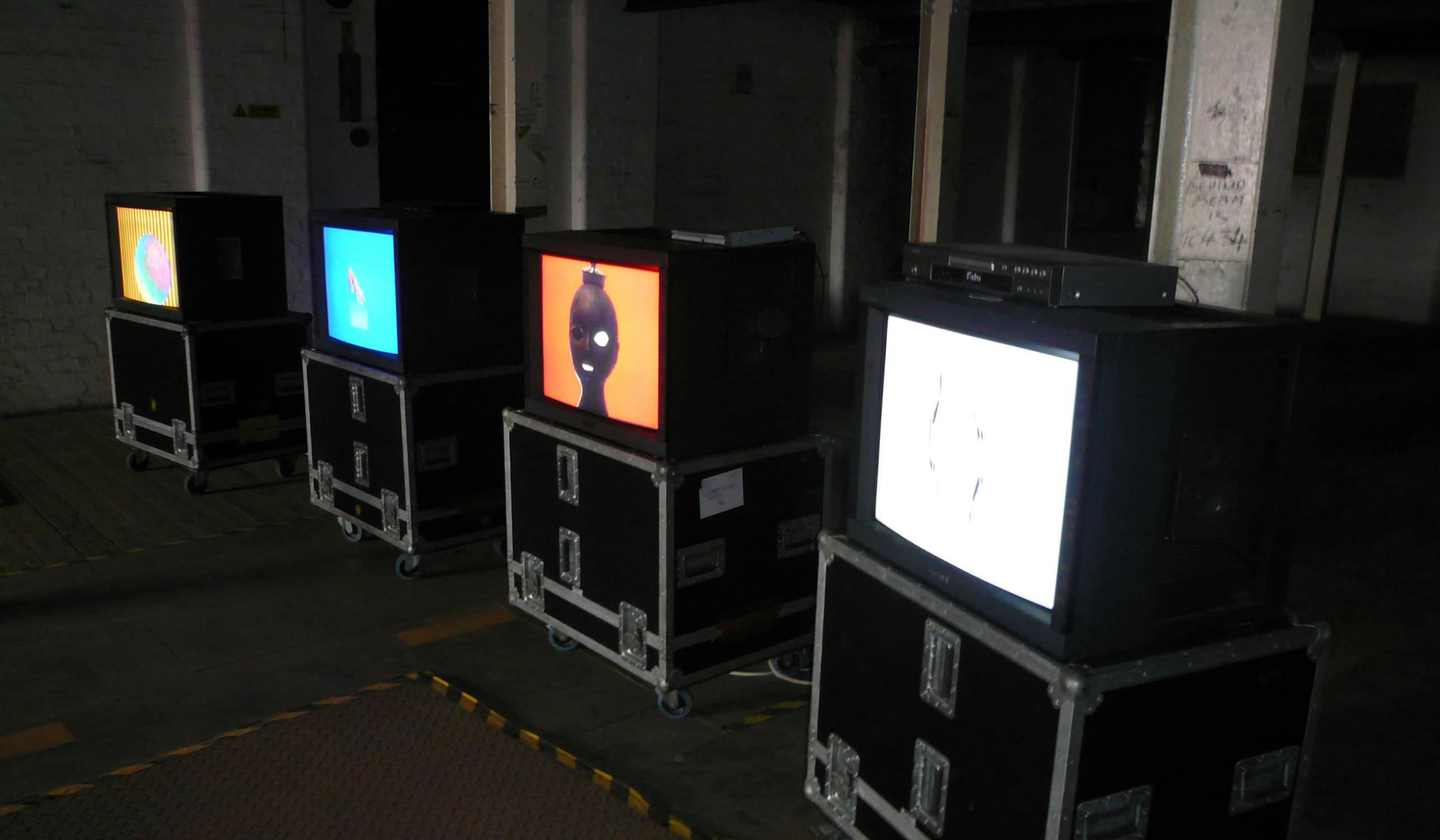 A row of old CRT televisions, each displaying different abstract images with vibrant colors, is set up on wheeled cases in a dimly lit, industrial-style space. The atmosphere is dark, and the equipment is arranged on a concrete floor.