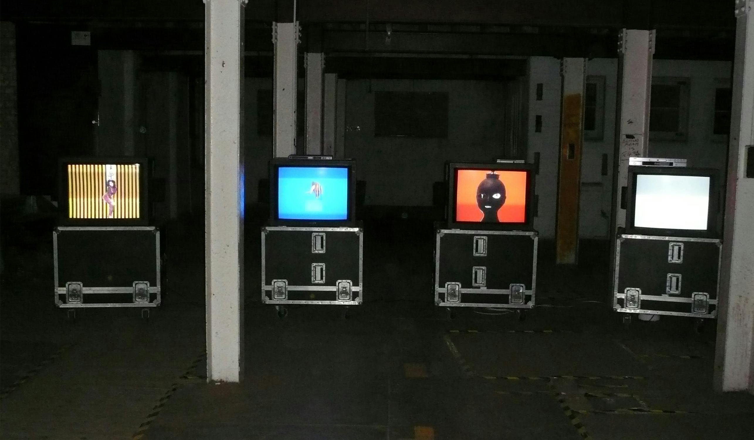 Four CRT televisions on metal stands display colorful graphics in a dimly lit industrial space. The screens show various abstract designs against a dark background.