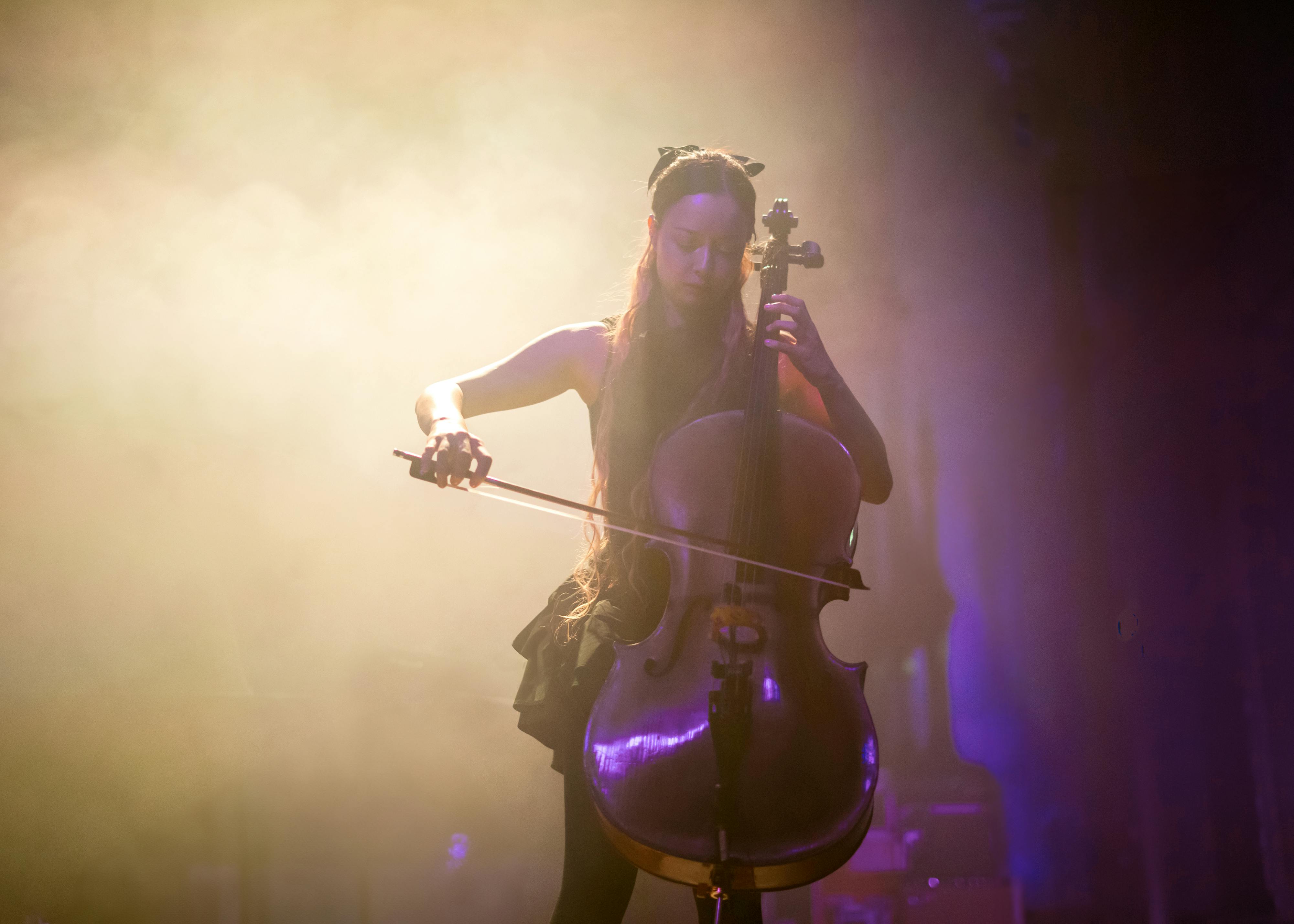 A person plays the cello on a dimly lit stage, surrounded by atmospheric fog. They are focused on the instrument, with long hair visible. The scene conveys a serene and emotive musical performance.