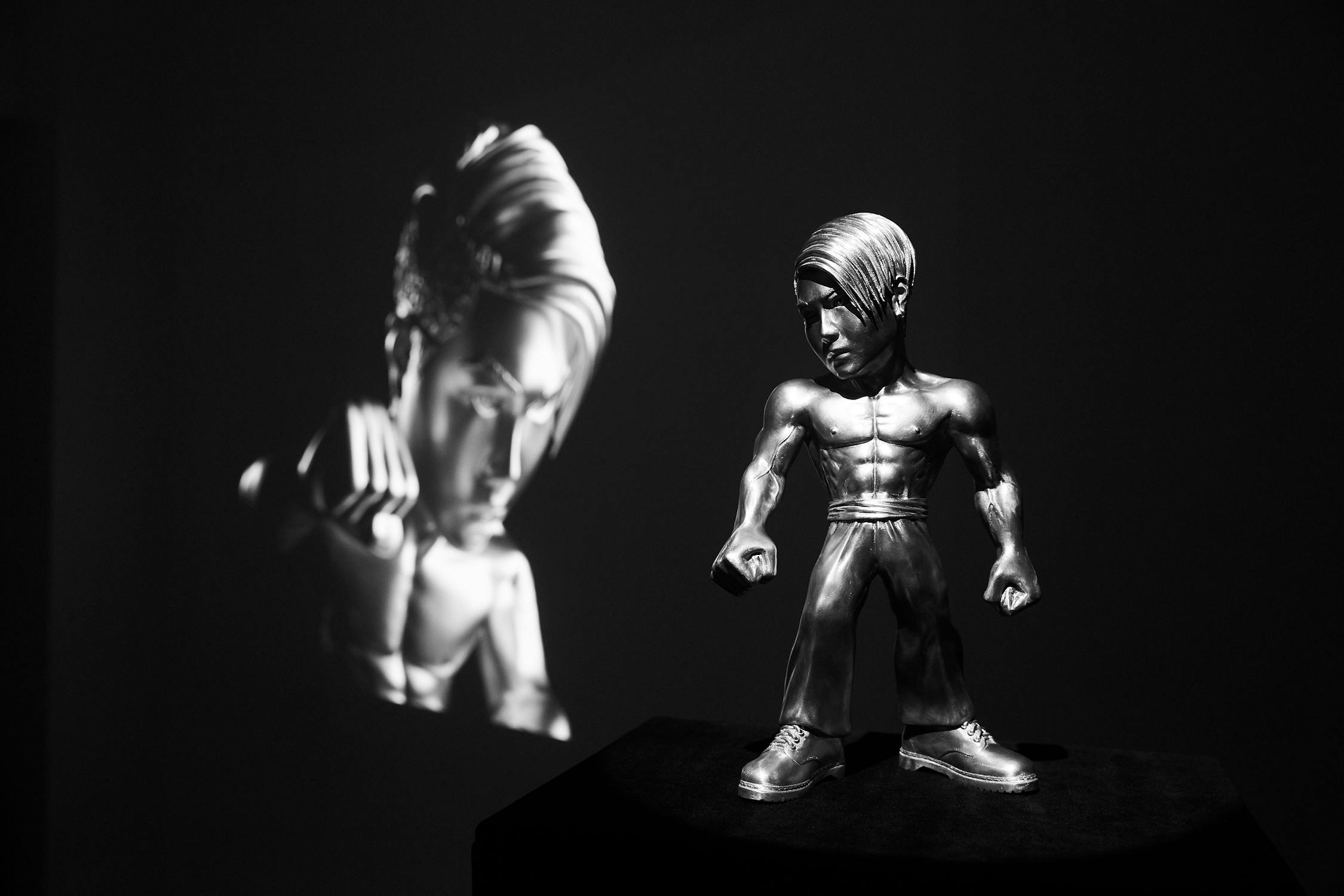 A metallic sculpted figure with defined muscles and styled hair stands on a platform. Its shadow is projected large on a wall behind, reflecting the same detailed features. The scene is lit dramatically in black and white.