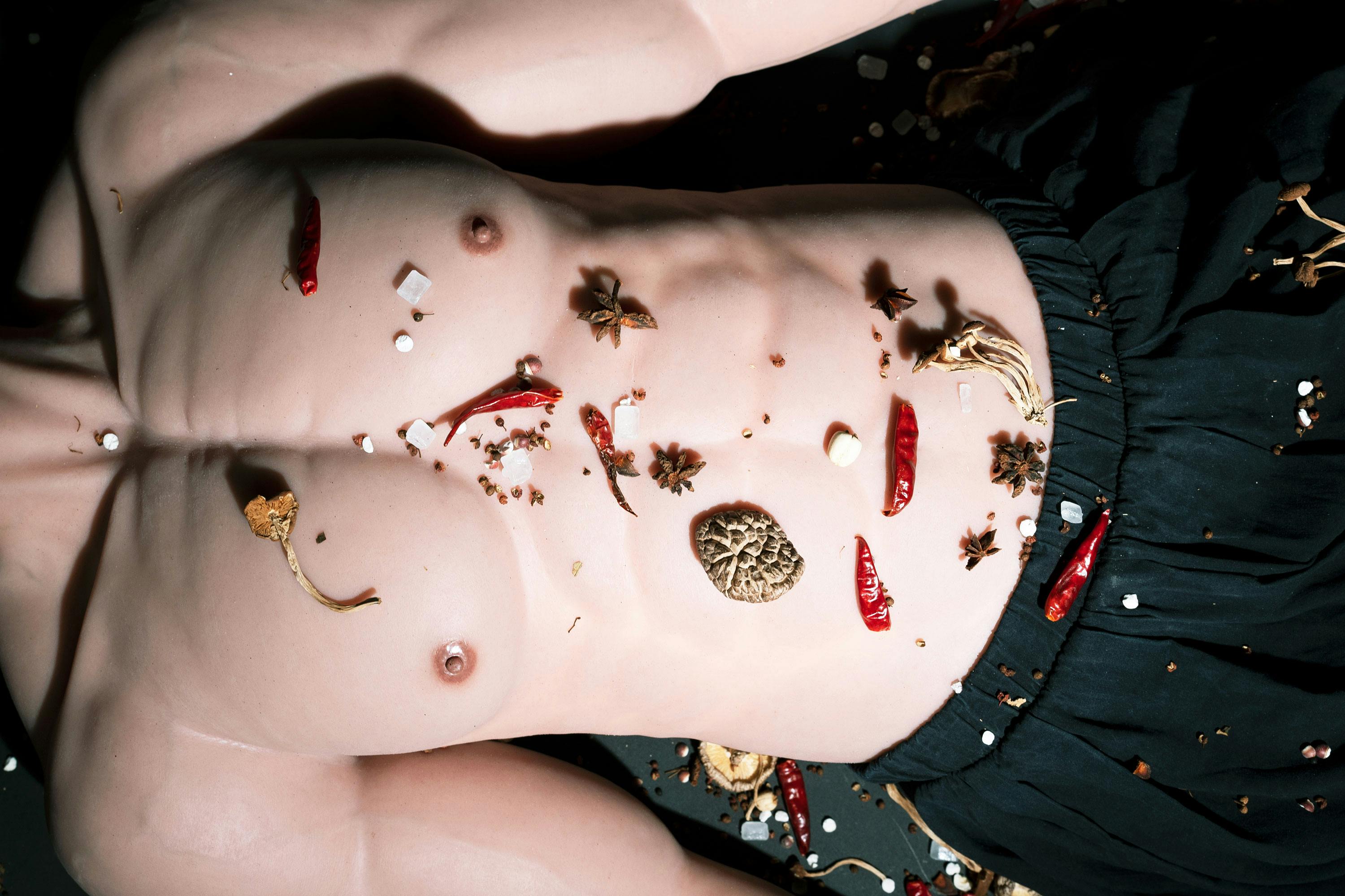 A close-up of a male torso with a bare chest and black pants, covered in various spices, including red chilli peppers, star anise, and dried herbs. The spices are scattered across the skin and surrounding area.