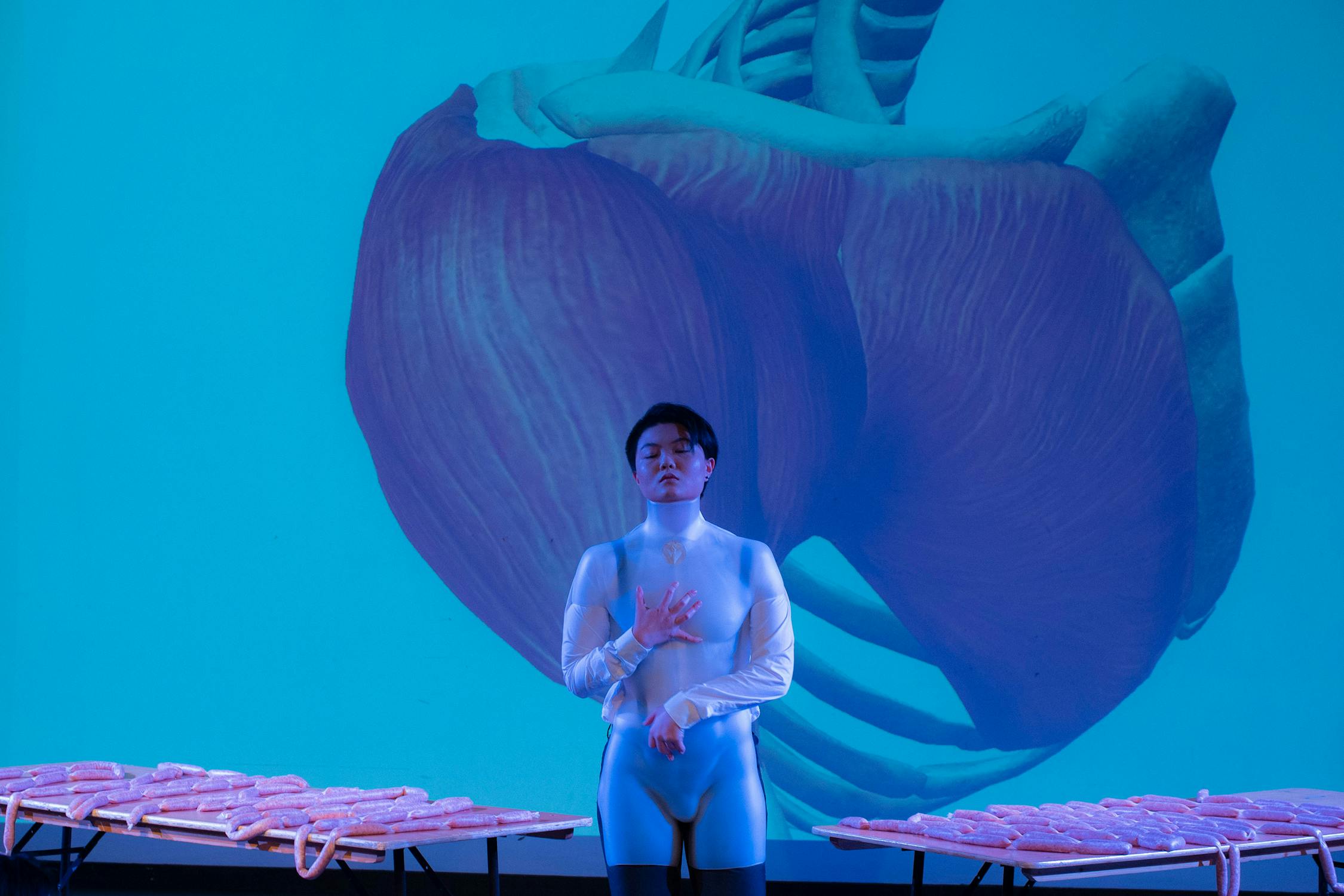 Whiskey Chow in a form-fitting outfit stands with eyes closed, hand on their chest, in front of a large anatomical image of a heart. The background is blue, and pink objects are laid out on tables on either side.