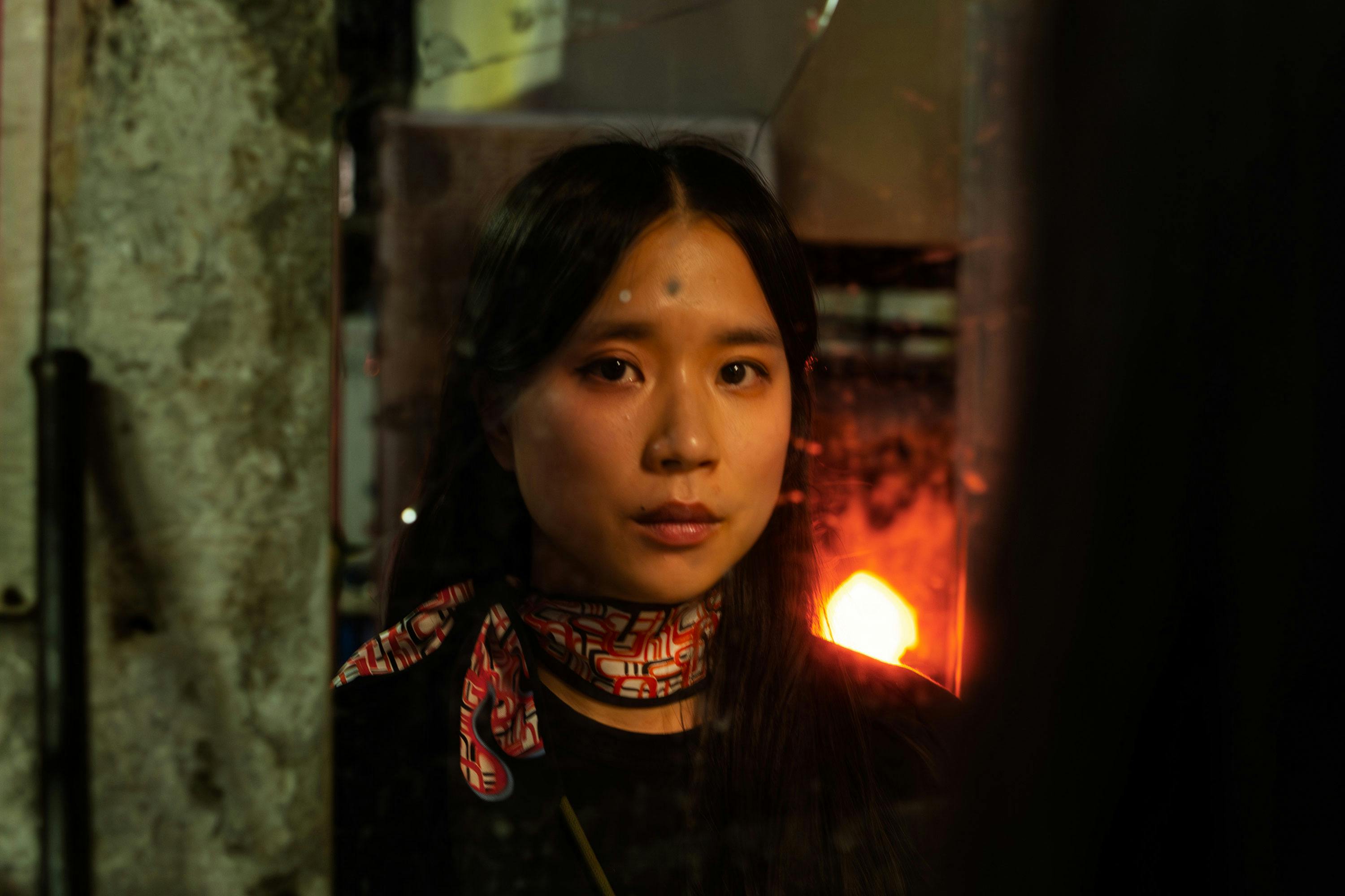 A woman with long dark hair is looking into a mirror, reflecting her serious expression. She wears a patterned scarf around her neck. The background has a warm, fiery glow, suggesting an industrial or workshop setting.