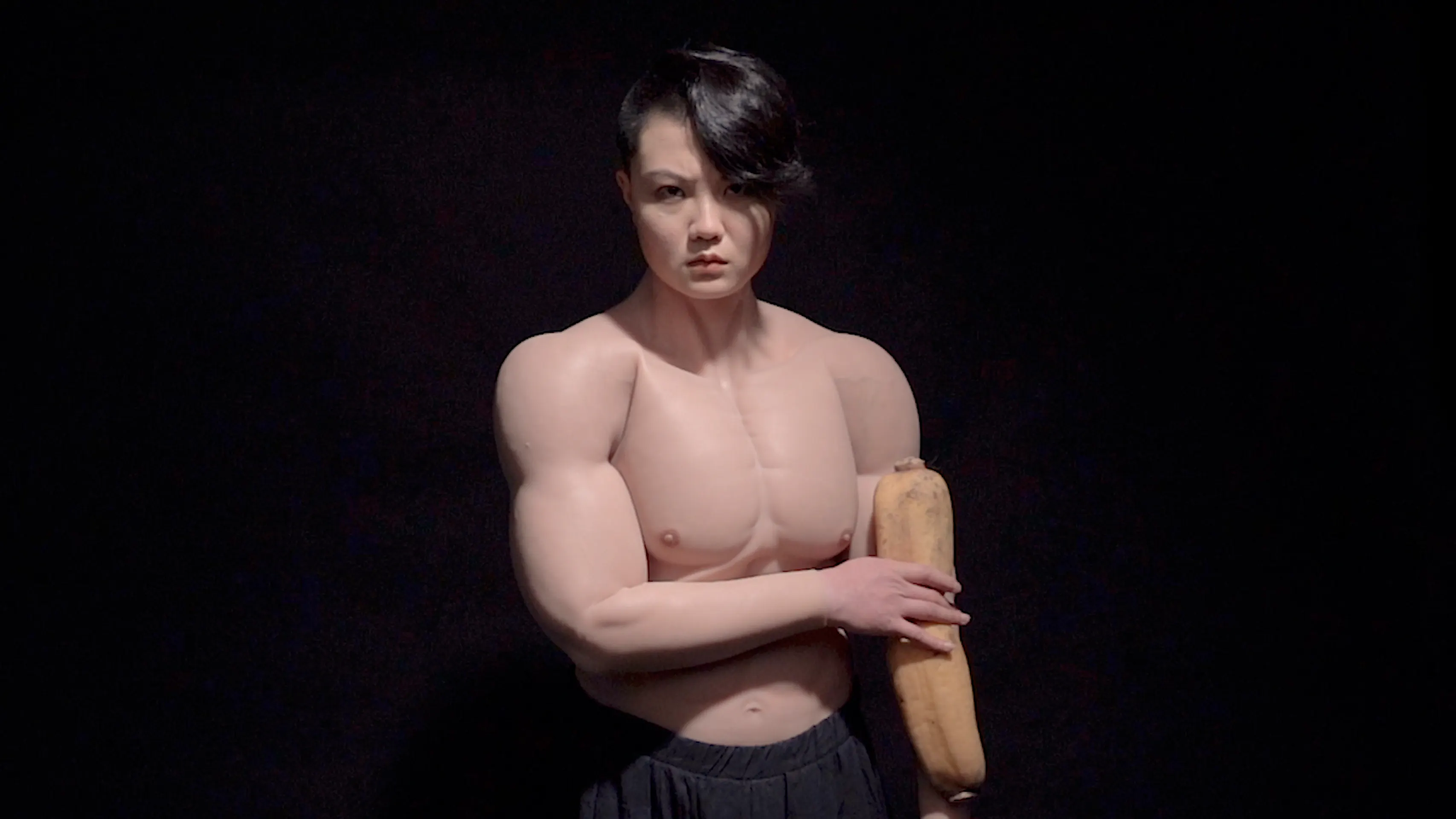 A muscular person with short, dark hair stands against a black background, holding a long loaf of bread. They are shirtless, highlighting their defined physique, and are wearing dark pants.