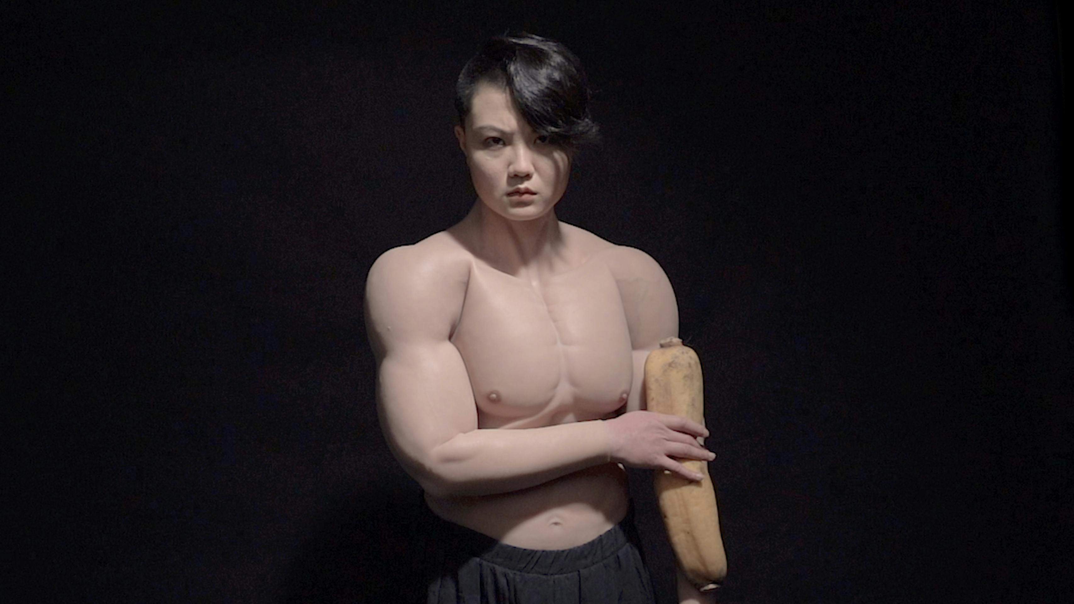 A muscular person with short, dark hair stands against a black background, holding a long loaf of bread. They are shirtless, highlighting their defined physique, and are wearing dark pants.