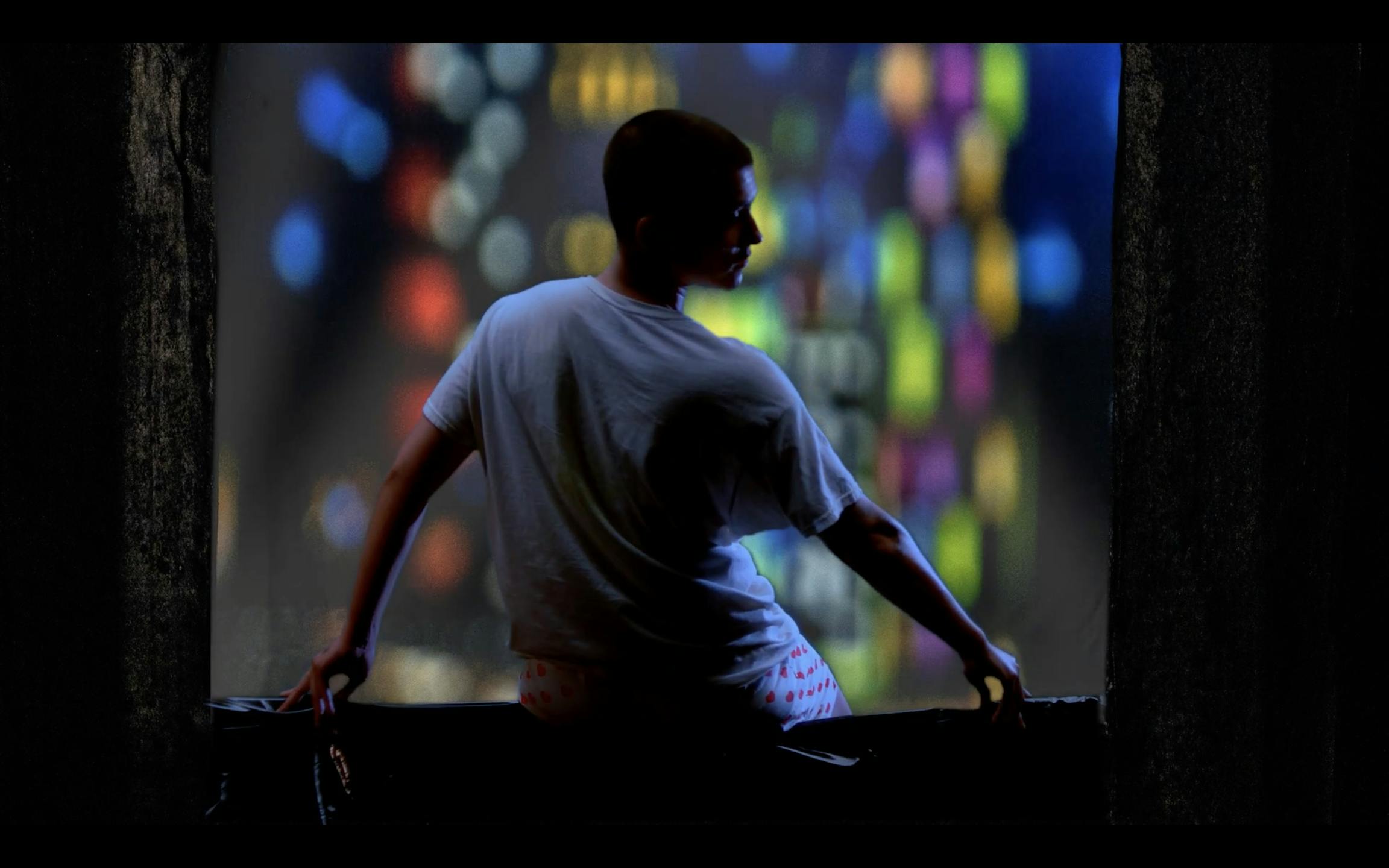 A person sits on a ledge, wearing a white T-shirt and shorts, with colorful city lights blurred in the background, creating a vibrant bokeh effect. The scene is dimly lit, emphasizing the contrast between the figure and the lights.