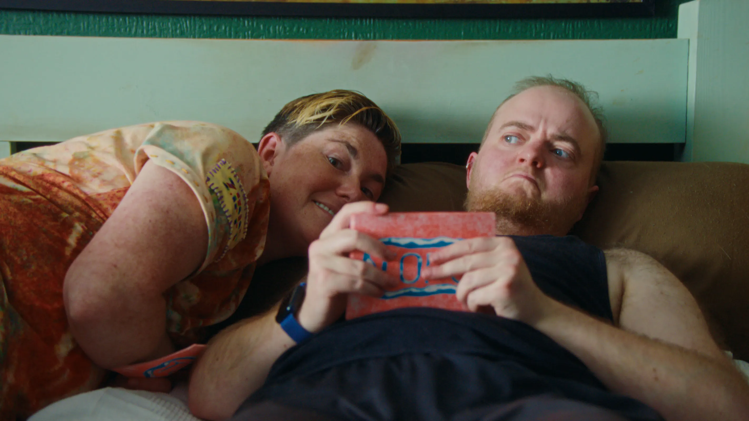 A person with short, two-toned hair smiles whilst leaning over a person with a beard who is looking down at a small, rectangular red card held in his hands. Both are lying in bed together.