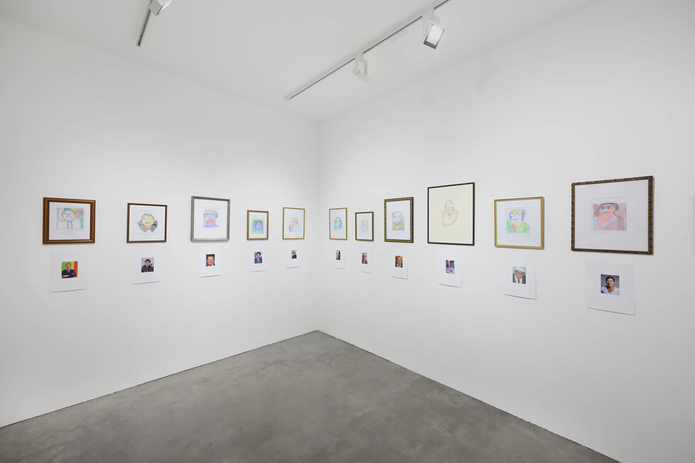 A white gallery wall displays framed portraits, each paired below with a smaller printed photo of a person. The floor is concrete, and the room is lit by track lighting. The arrangement suggests a comparison between the drawings and colour photos.