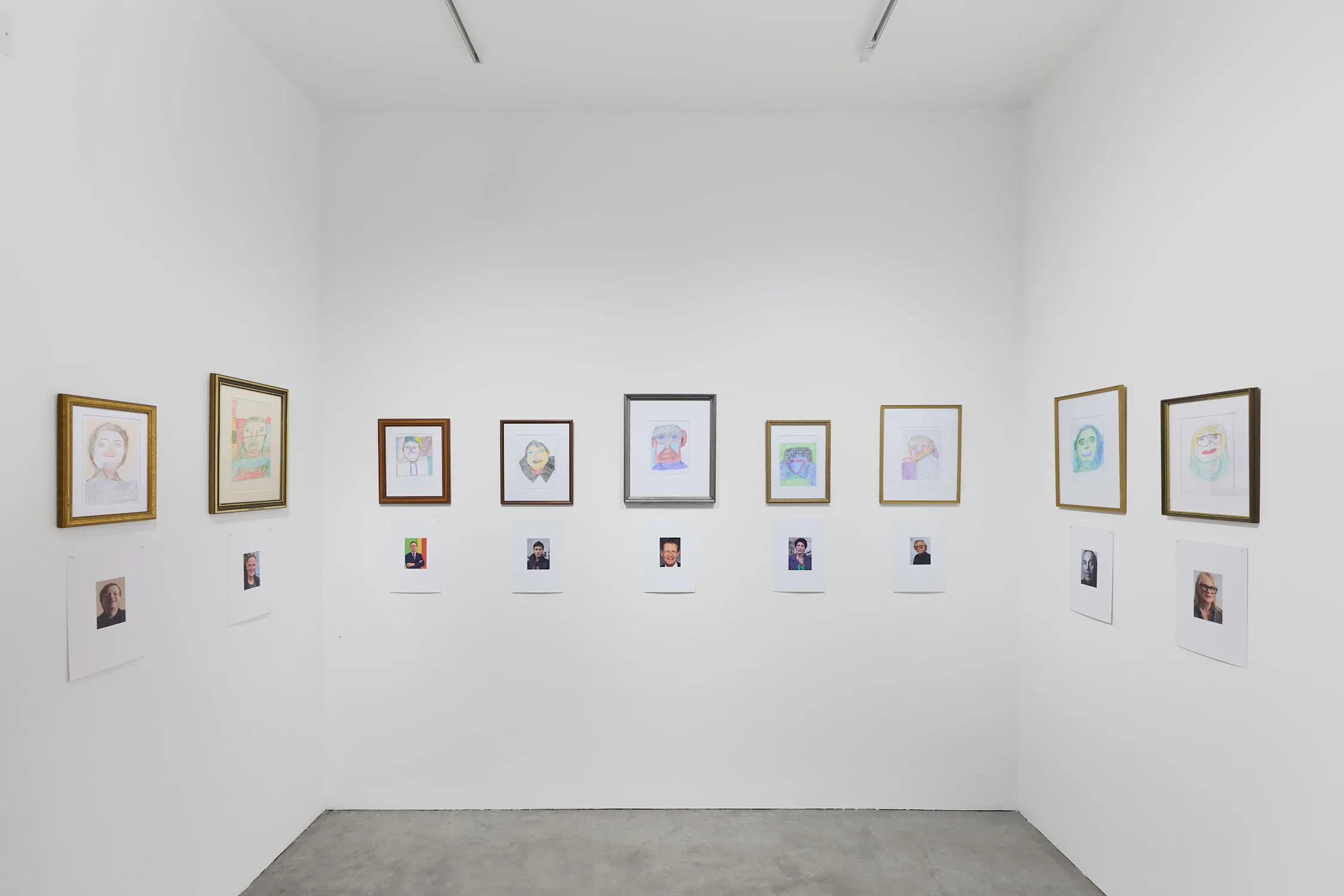 A small art gallery room displays framed hand-drawn portraits on white walls, each hung above a photo of a person's face. The floor is grey concrete, and the space is lit by overhead lights. The arrangement creates a formal, symmetrical presentation.