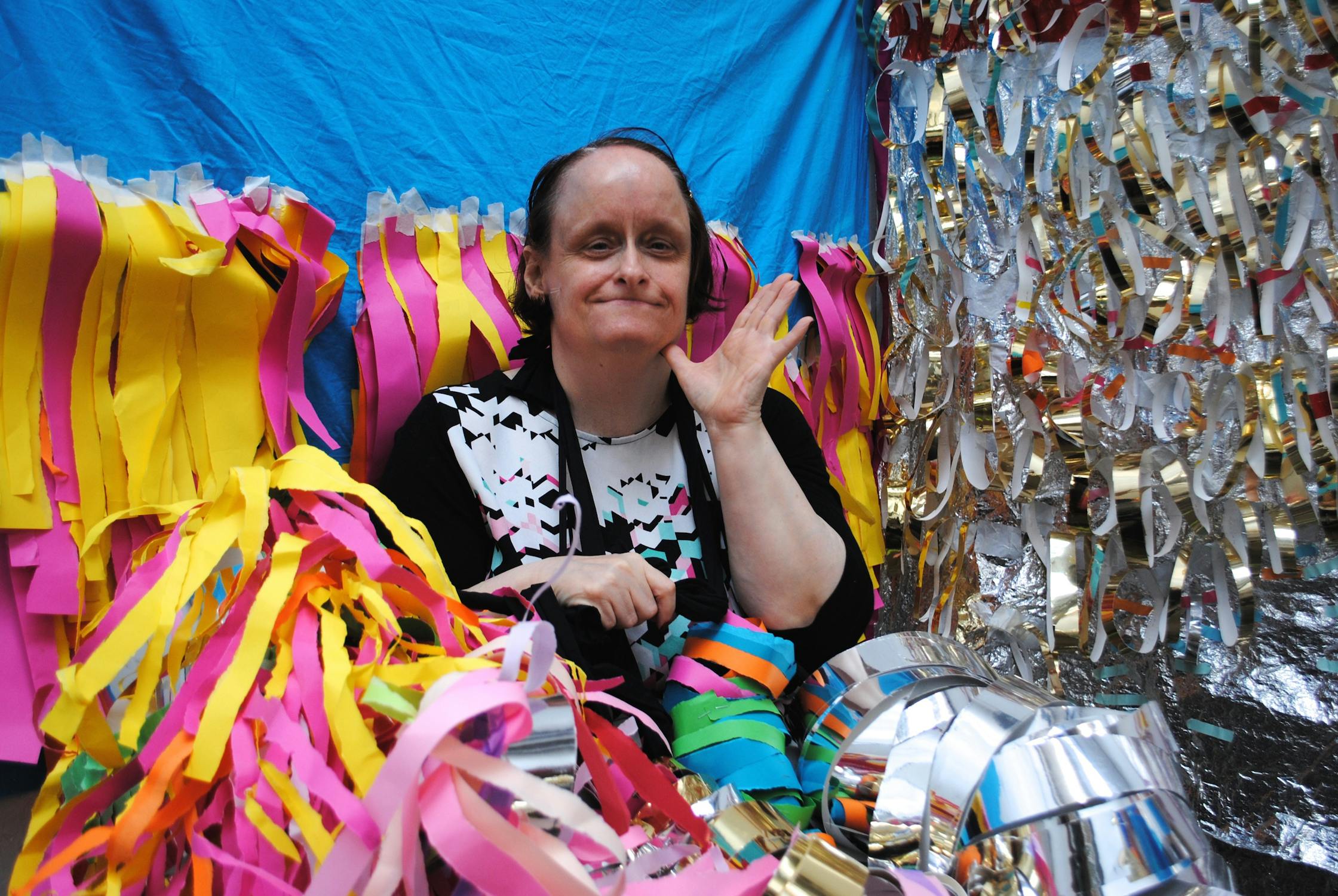 A person smiling and waving, surrounded by colorful streamers and foil decorations. Their backdrop is a mix of shiny materials and a blue fabric.