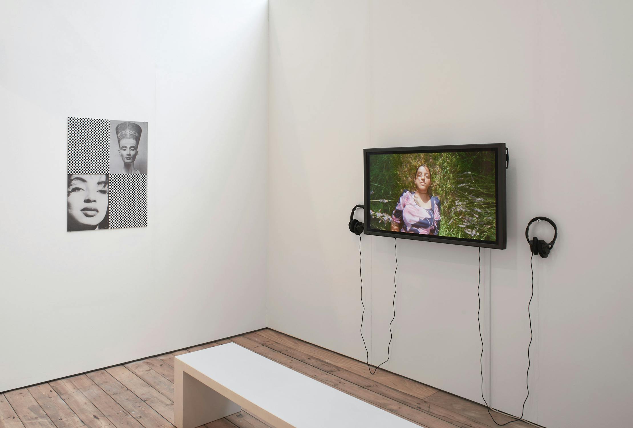A minimalist exhibition room features a black-and-white portrait on the wall and a video screen displaying a person in nature. Headphones hang beside the screen. A simple wooden bench is positioned on the wooden floor for viewing.