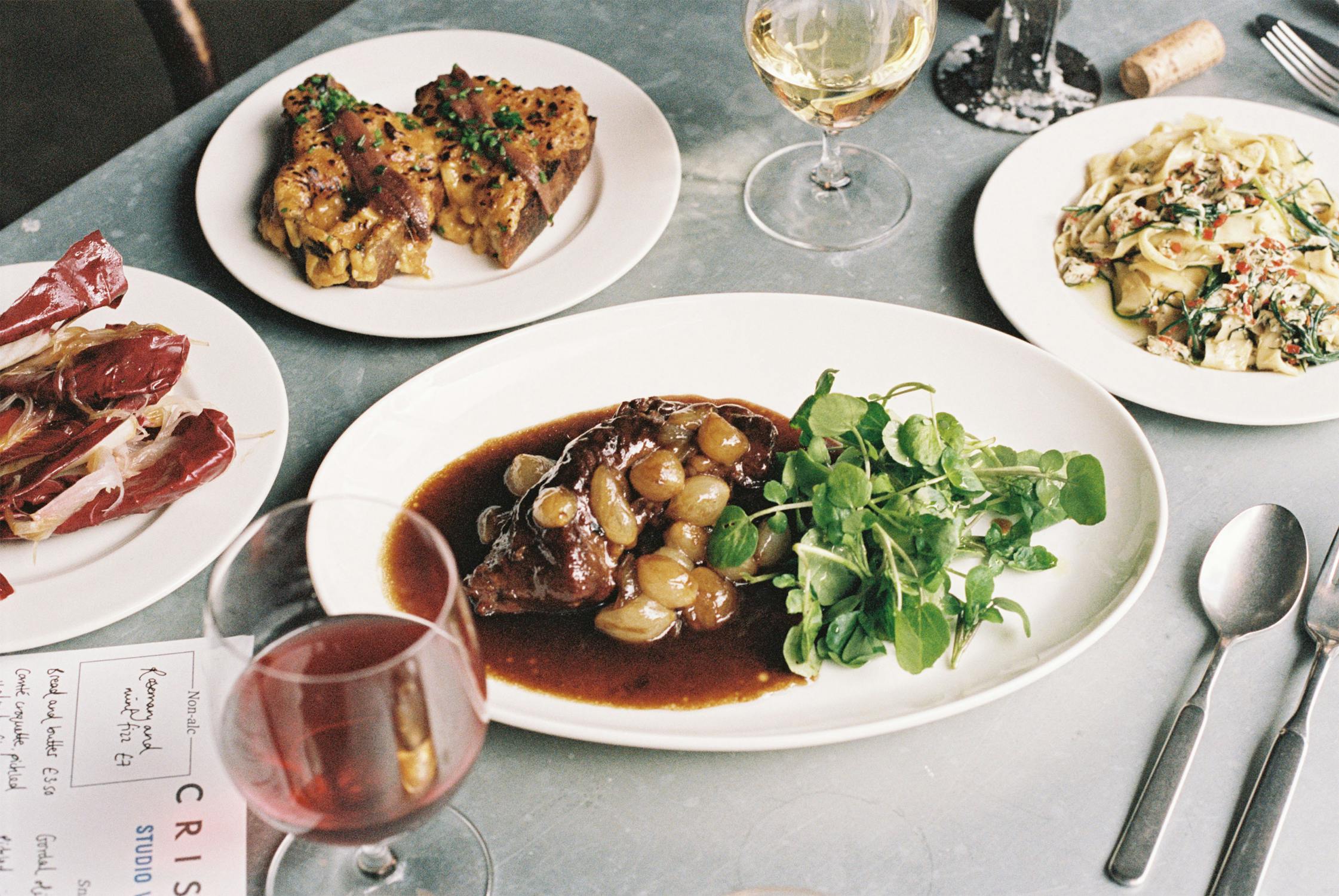 A photograph of a table with plates of food and drinks. In the centre there is a large serving plate with a steak and red wine sauce. There is a dressed salad, a plate of pasta. In the forground there is a large glass of red wine.