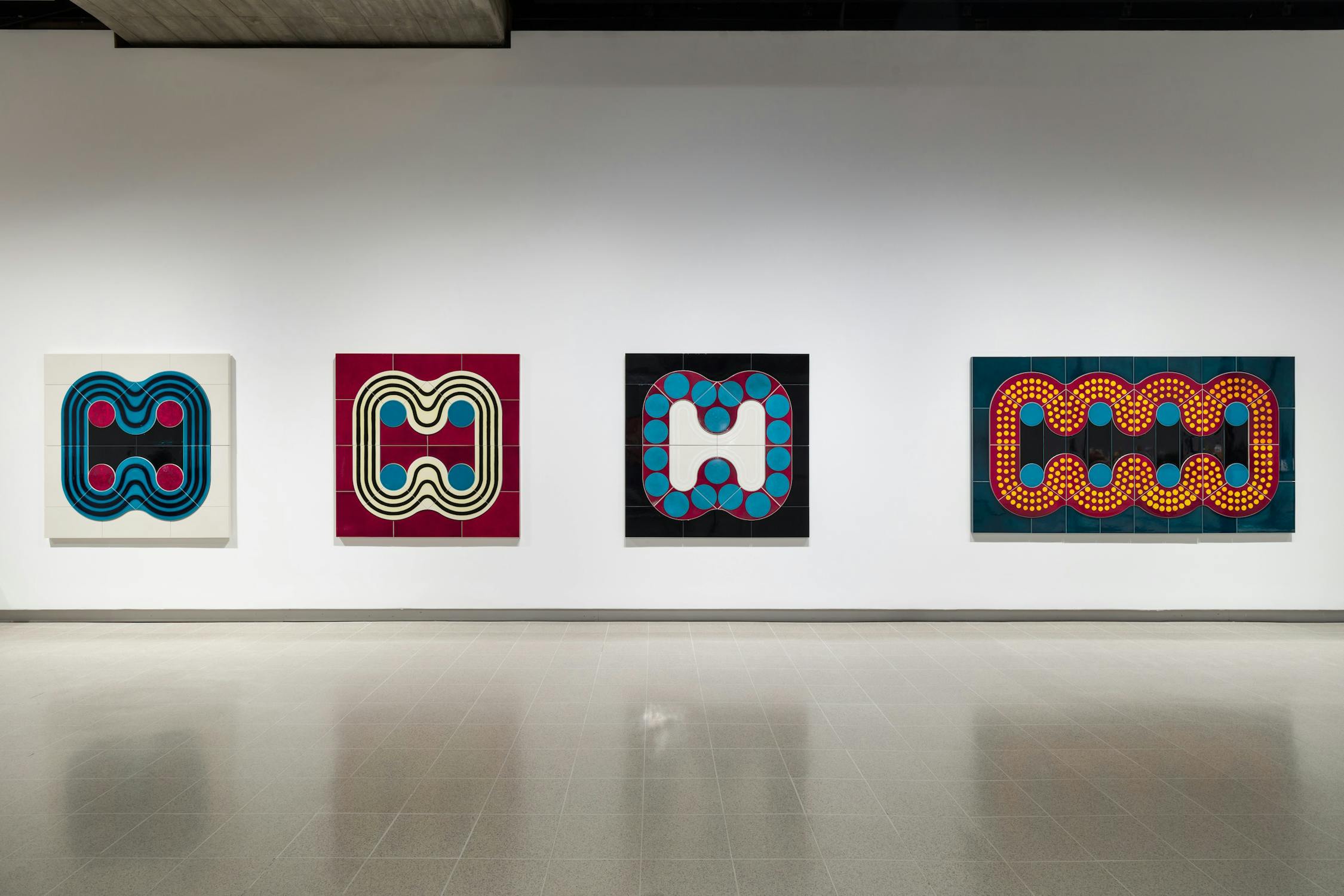 Four abstract paintings hang on a gallery wall. Each artwork features geometric patterns with vibrant colors, predominantly using circles and curving lines against contrasting backgrounds. The floor and ceiling elements are visible, enhancing the exhibit's presentation.