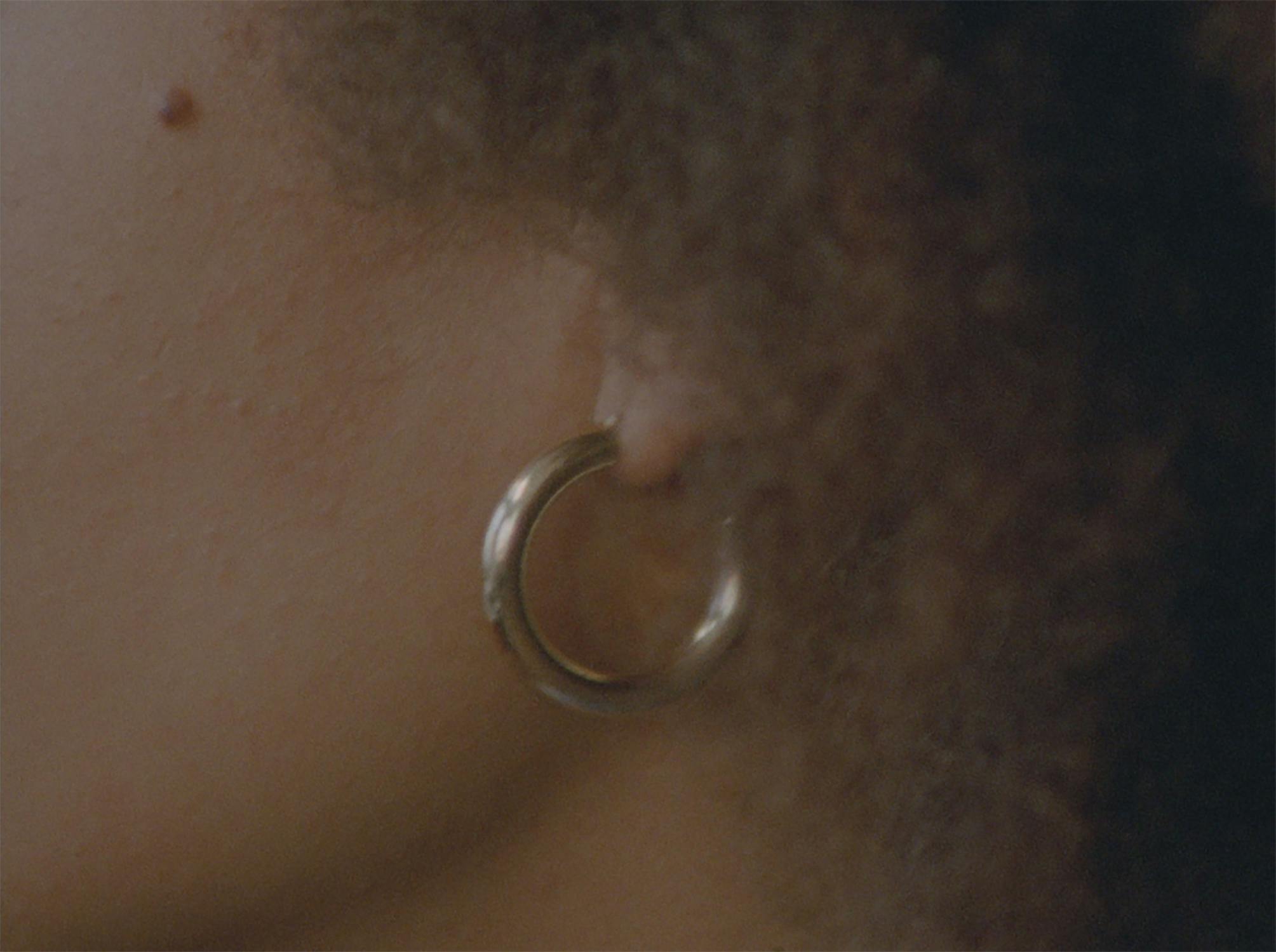 Close-up of a person's ear wearing a gold hoop earring. The person's curly hair partially covers the ear, and the skin is lightly freckled. The lighting is soft, creating a warm, intimate feel.