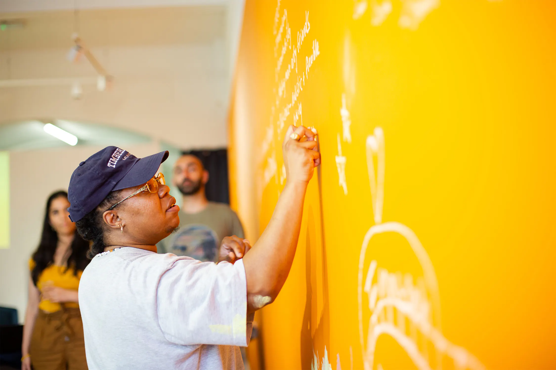 Exodus Crooks wearing glasses and a cap writes on a bright orange wall with white chalk, while two people stand in the background watching.