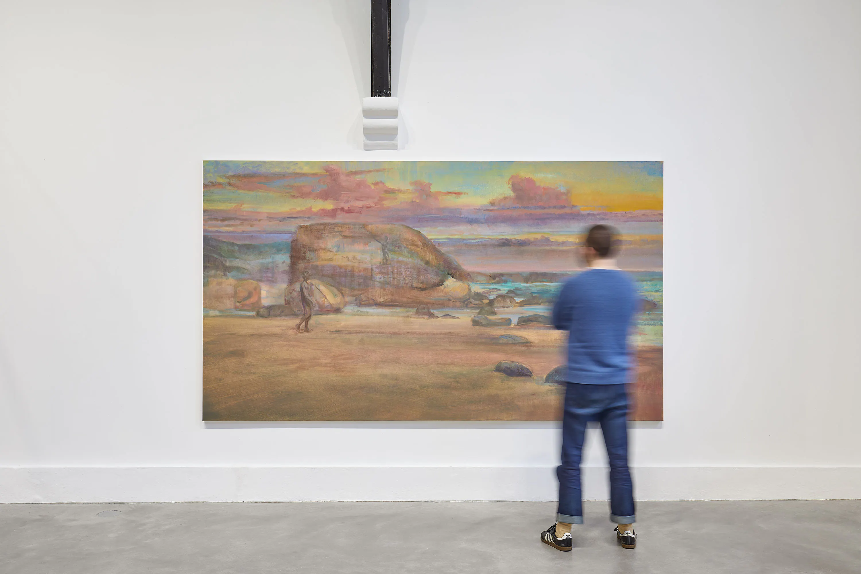 A person in a blue sweater and jeans stands in front of a large, colourful painting of a rocky beach scene displayed on a white gallery wall. The person's face and figure are slightly blurred.