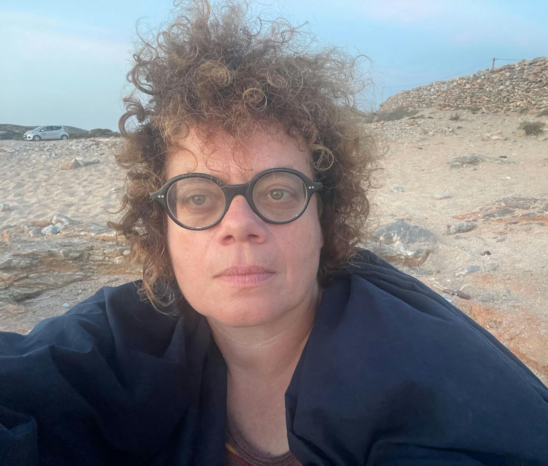 Portrait of Oreet Ashery seated on a rocky beach, wearing round glasses and a dark jacket, with curly hair and a calm expression.
