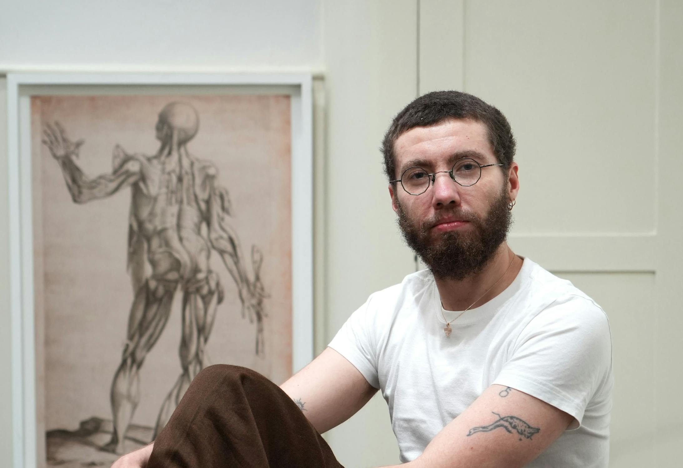 Portrait of Caspar Heinemann seated indoors, wearing round glasses, a white t-shirt, and brown trousers, with visible tattoos and an anatomical drawing in the background.