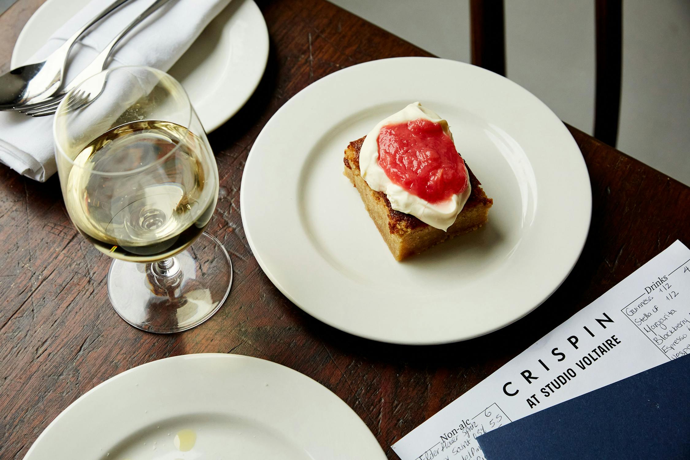 A glass of white wine and a dessert with cream and pink topping on a white plate, placed on a wooden table next to cutlery and a printed menu labelled “CRISPIN AT STUDIO VOLTAIRE”.